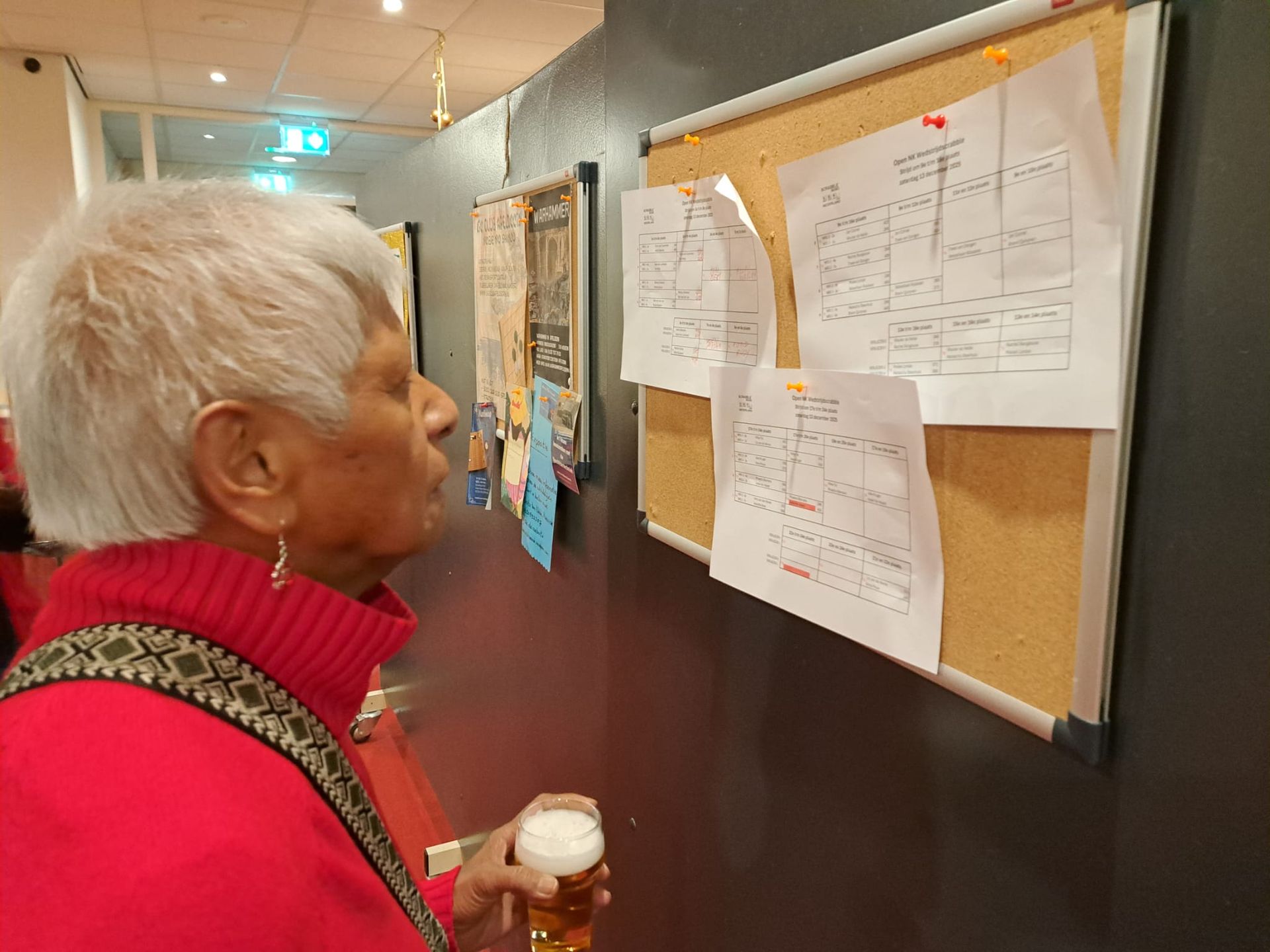 Een persoon bekijkt papieren die op een prikbord aan de muur zijn vastgeprikt, terwijl hij een glas bier vasthoudt.