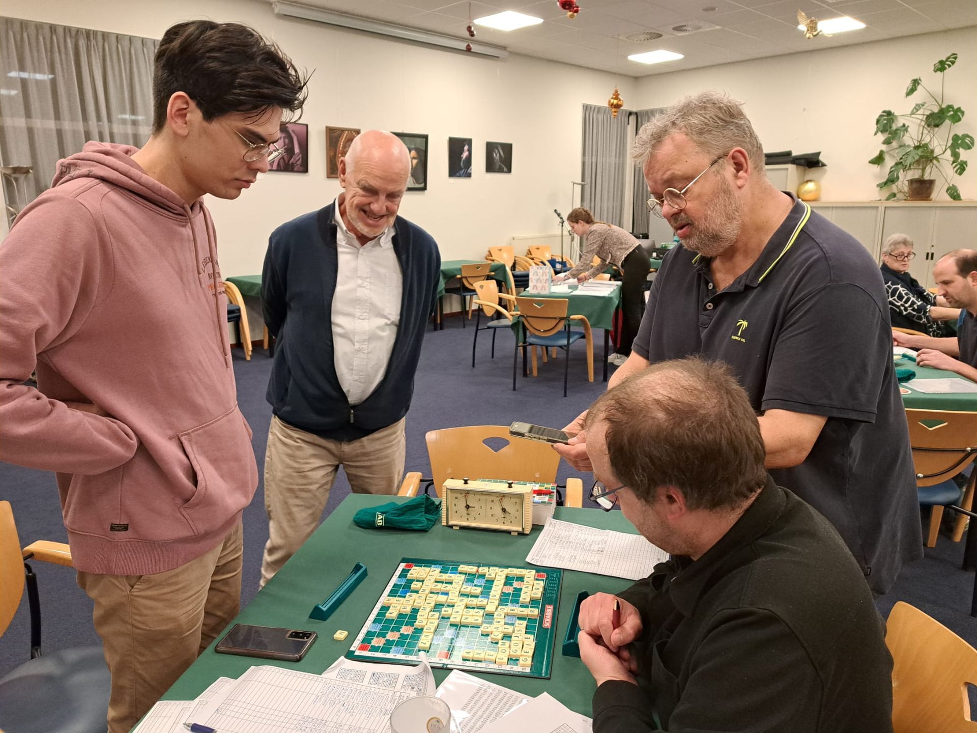 Vier mannen zitten rond een bordspel. Een van hen gebaart en legt het spel uit. Anderen kijken aandachtig toe in een kamer met tafels.