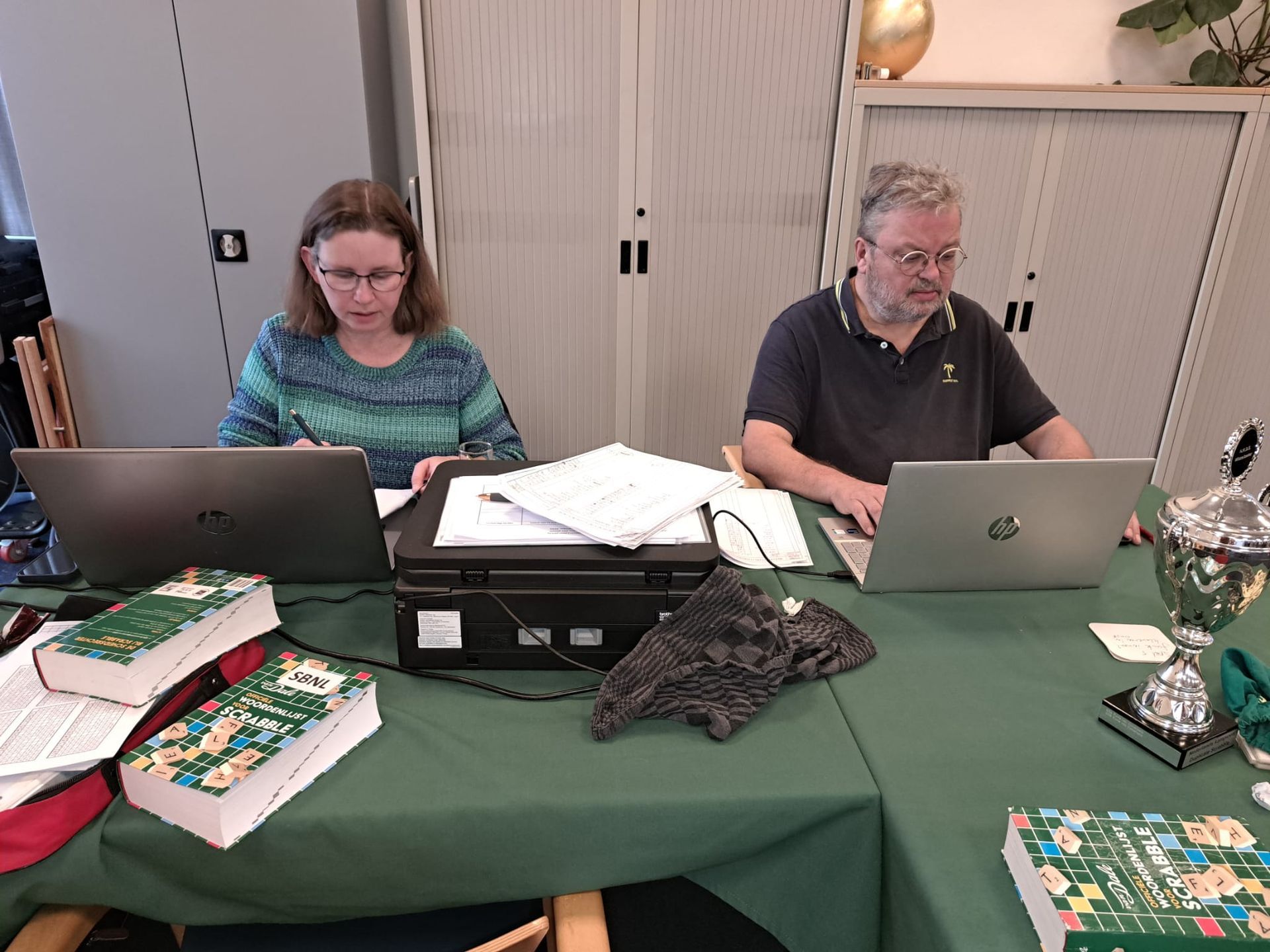Twee mensen werken aan een tafel met laptops, een printer, boeken en een trofee, binnenshuis.