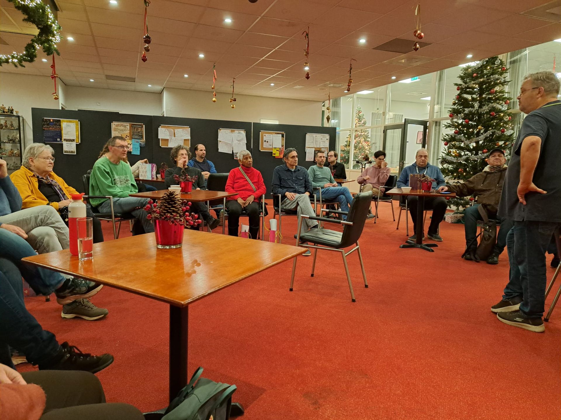 Een groep mensen zit aan tafels en luistert naar een spreker. Binnen ligt een rode loper en er staat een kerstboom.