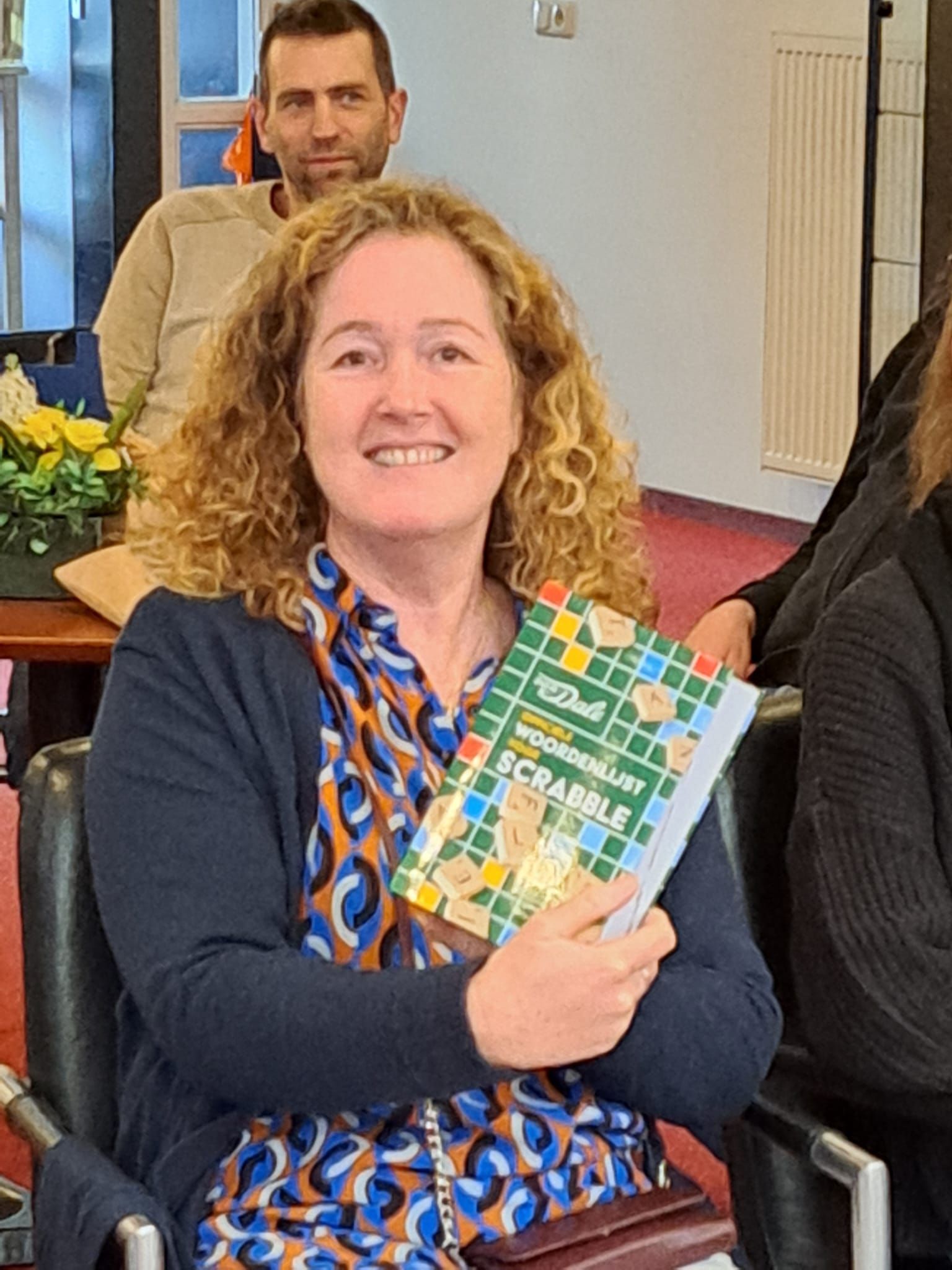 Vrouw met krullend haar glimlacht, terwijl ze een Scrabble-boek vasthoudt, zittend in een kamer. Een man is op de achtergrond te zien.