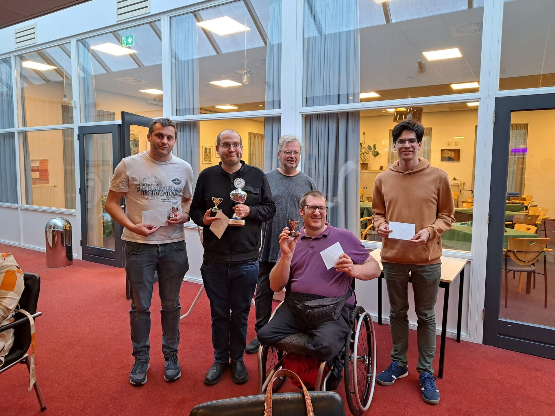 Vijf mensen poseren met trofeeën/certificaten in de hand, vlakbij glazen deuren/ramen, en glimlachen. Eén van hen zit in een rolstoel.