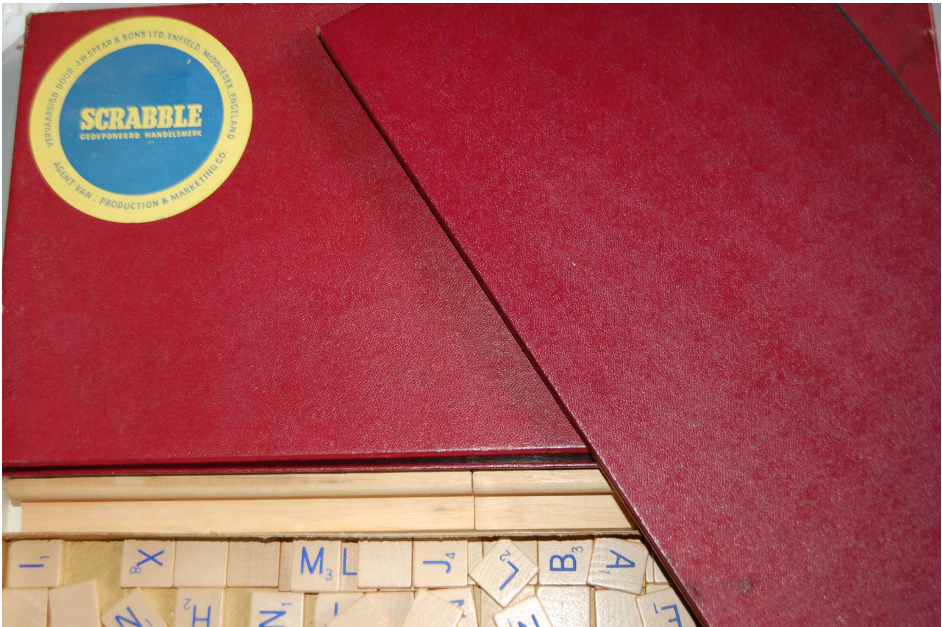 Rode Scrabble-doos, gedeeltelijk open, met houten letterblokjes erin. Blauwe Scrabble-sticker op de bovenkant.