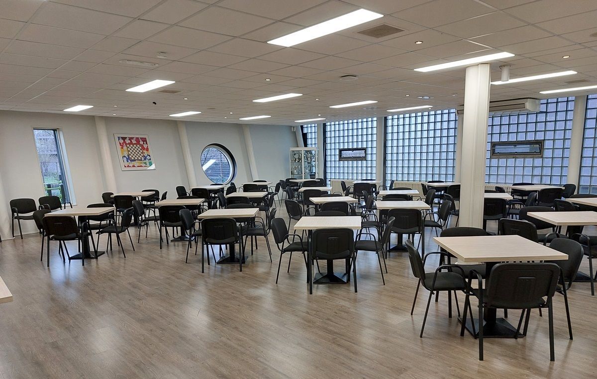 Lege kantine met tafels, stoelen en grote ramen. Grijze vloer, witte muren en plafond.