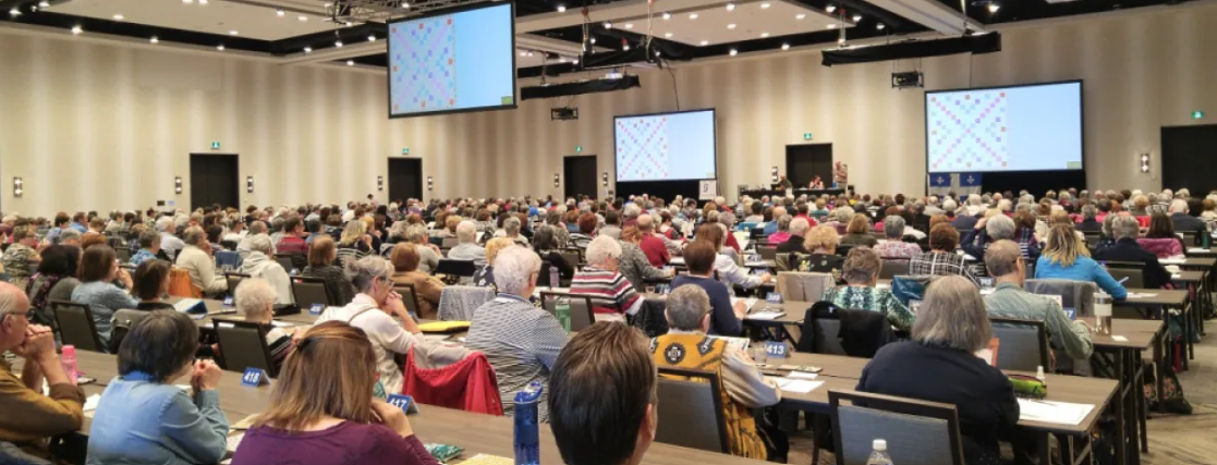 Grote conferentiezaal met honderden mensen die aan tafels zitten, tegenover een podium met schermen.