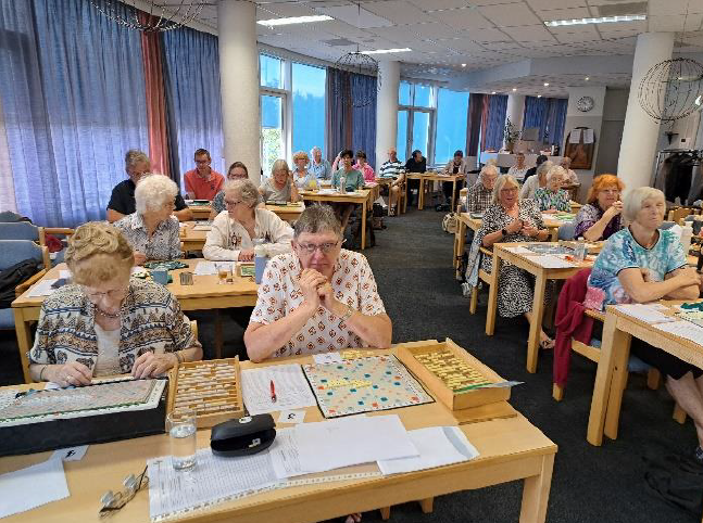 Mensen spelen een bordspel aan tafels in een felverlichte ruimte. Sommigen zijn geconcentreerd, anderen kijken toe.