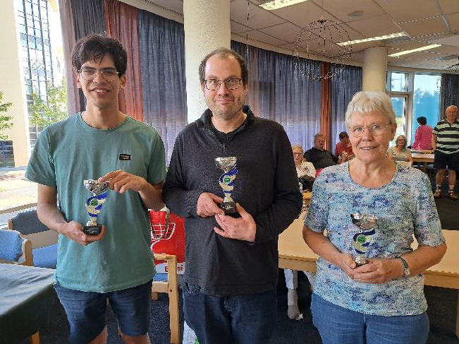 Drie mensen met trofeeën; binnen. Links: Jongeman in turquoise shirt lacht. Midden: Man met bril. Rechts: Oudere vrouw in blauw shirt lacht.