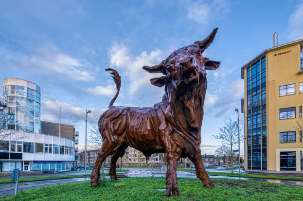 Een groot bronzen stierbeeld op een plein met gebouwen en een blauwe lucht op de achtergrond.