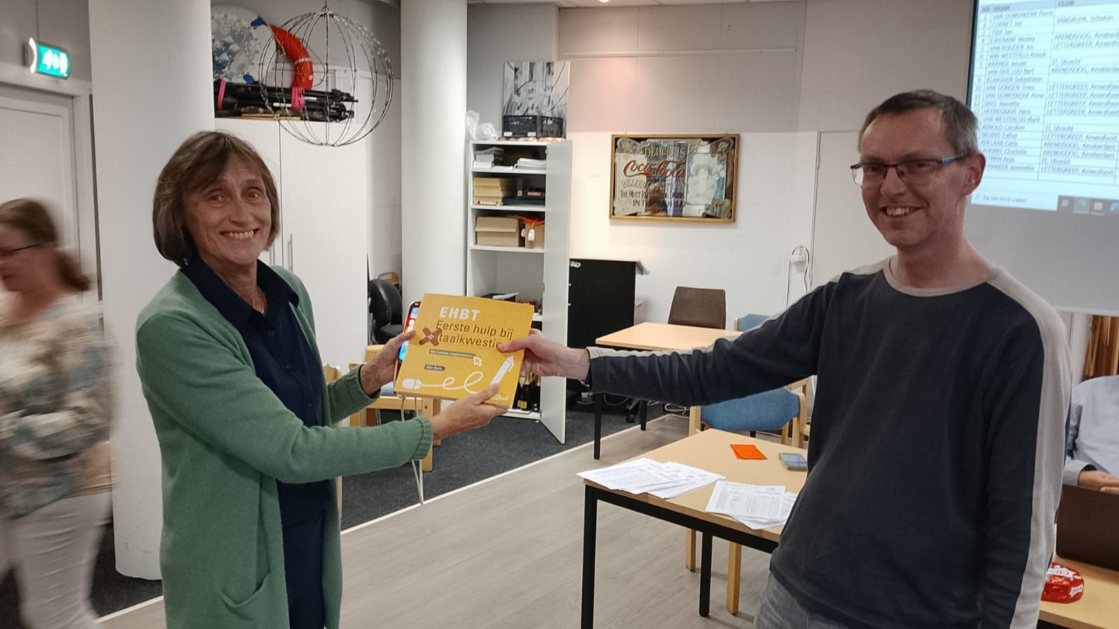 Een vrouw in een groene jas overhandigt een boek aan een man met een bril in een kantooromgeving.