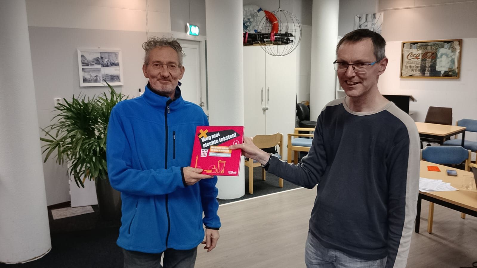 Twee mannen houden een boek met een rode kaft vast en wijzen ernaar. Ze bevinden zich in een gebouw met een plant en kunstwerken.