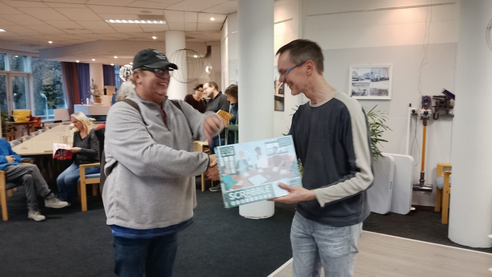 Een man met een hoed overhandigt een cadeau aan een andere man binnenshuis, met een glimlach.