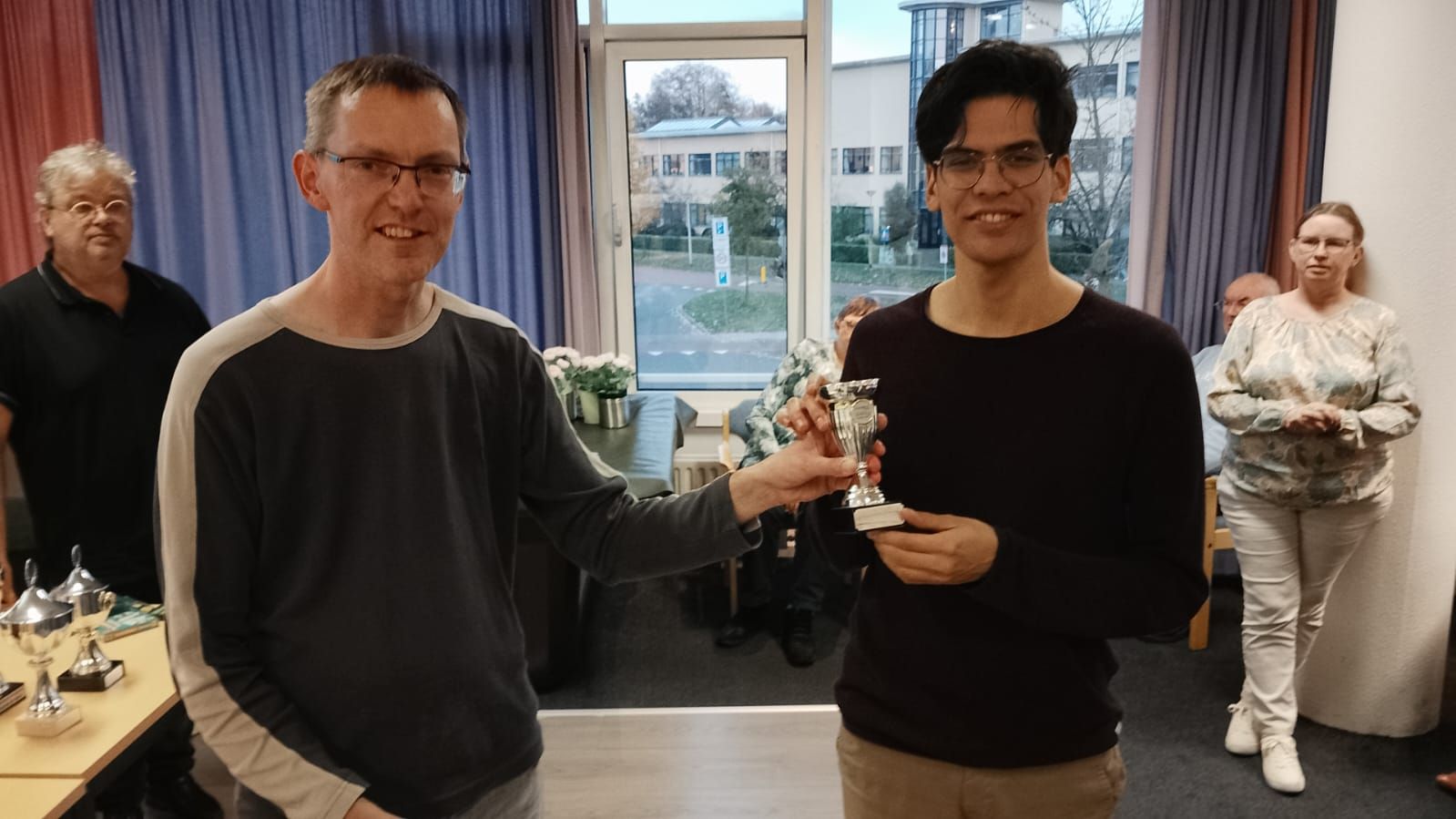 Twee mannen, van wie er één een trofee vasthoudt, glimlachen. Anderen kijken toe vanuit een kamer.