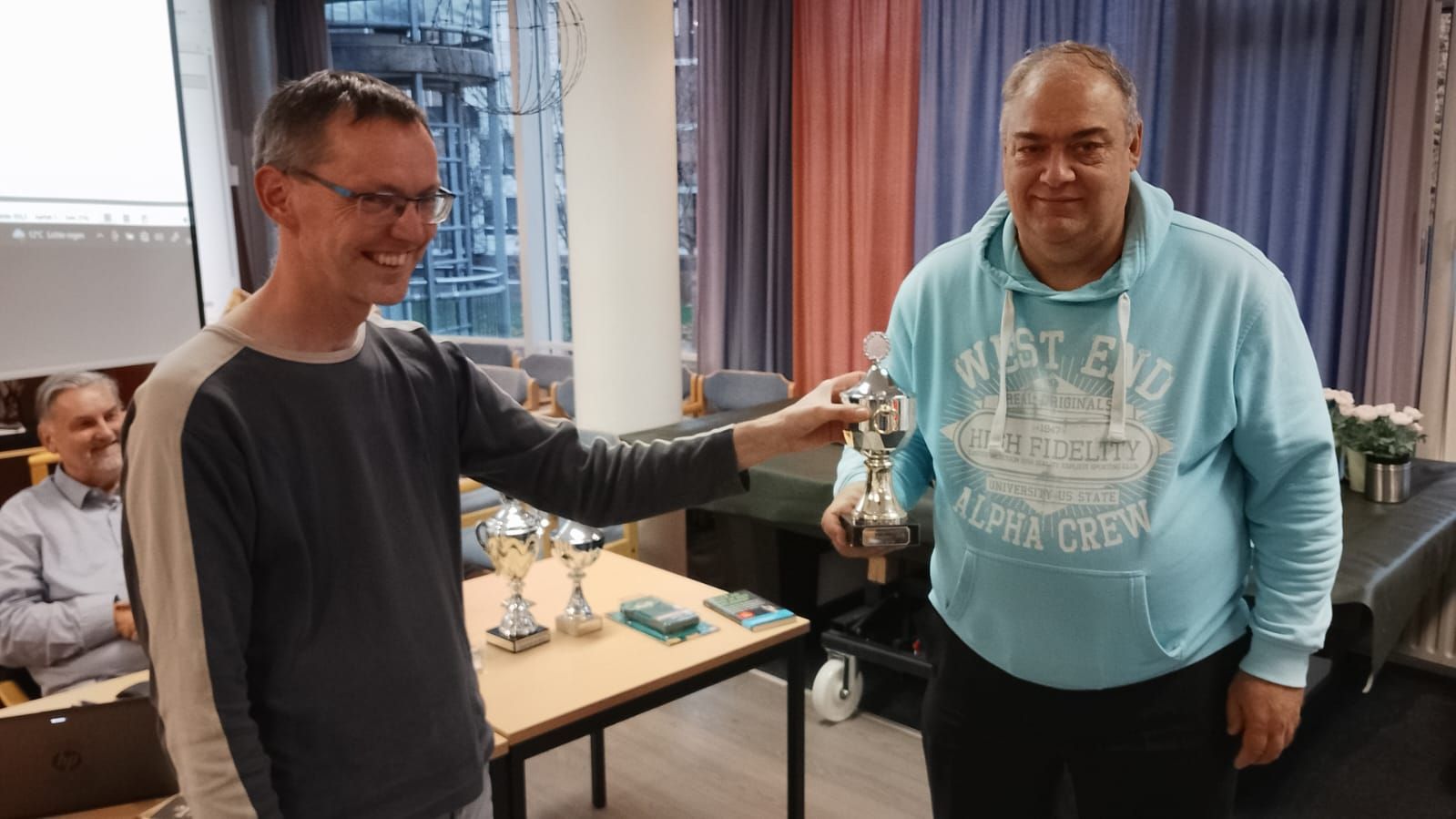 Een man overhandigt een trofee aan een andere man; beiden glimlachen. Op een tafel staan ​​andere trofeeën; op de achtergrond is een zittende man te zien.