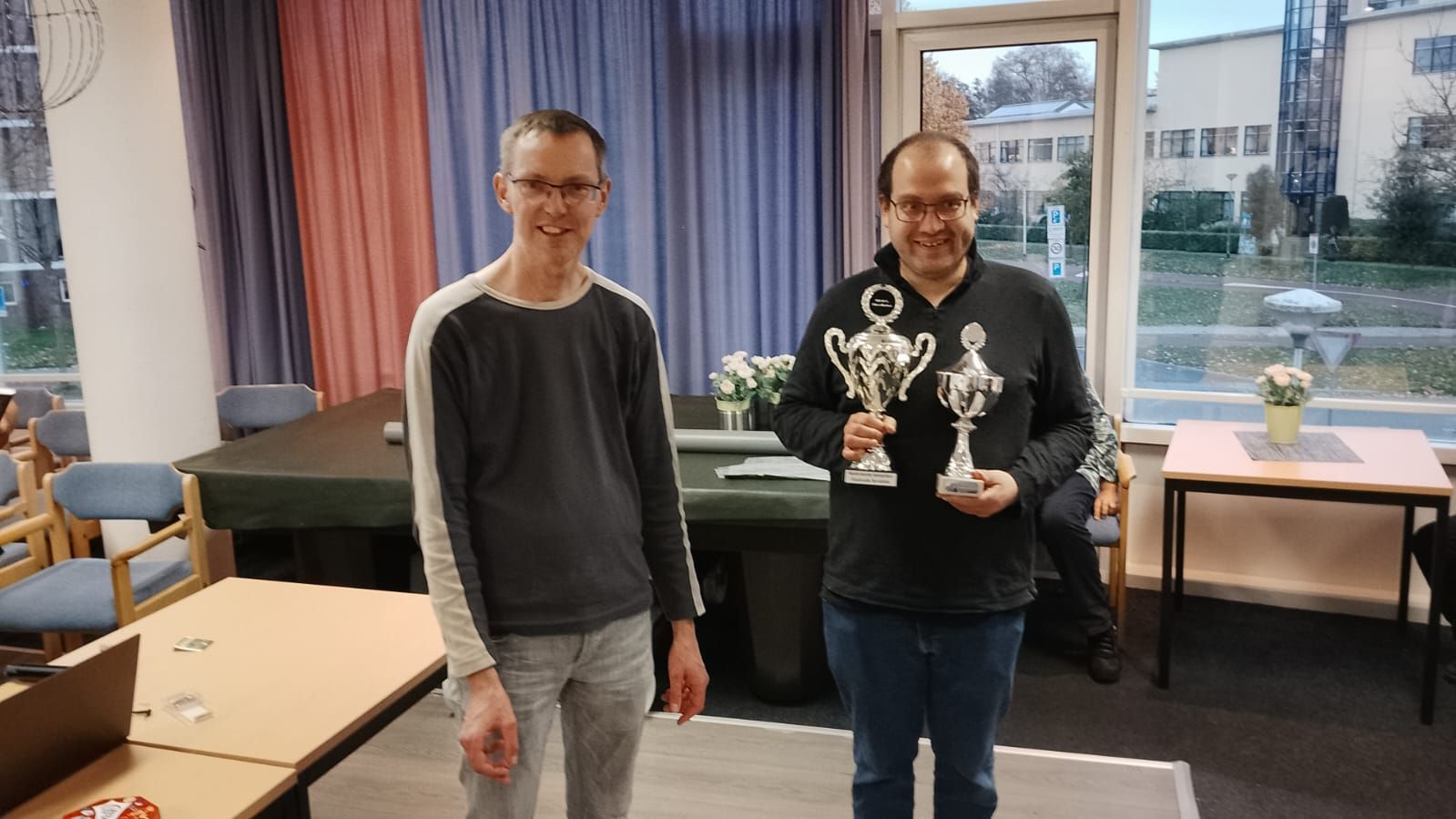 Twee mannen in een kamer met trofeeën in hun handen. De ene glimlacht, de andere kijkt neutraal. Neutraal interieur met een raam op de achtergrond.