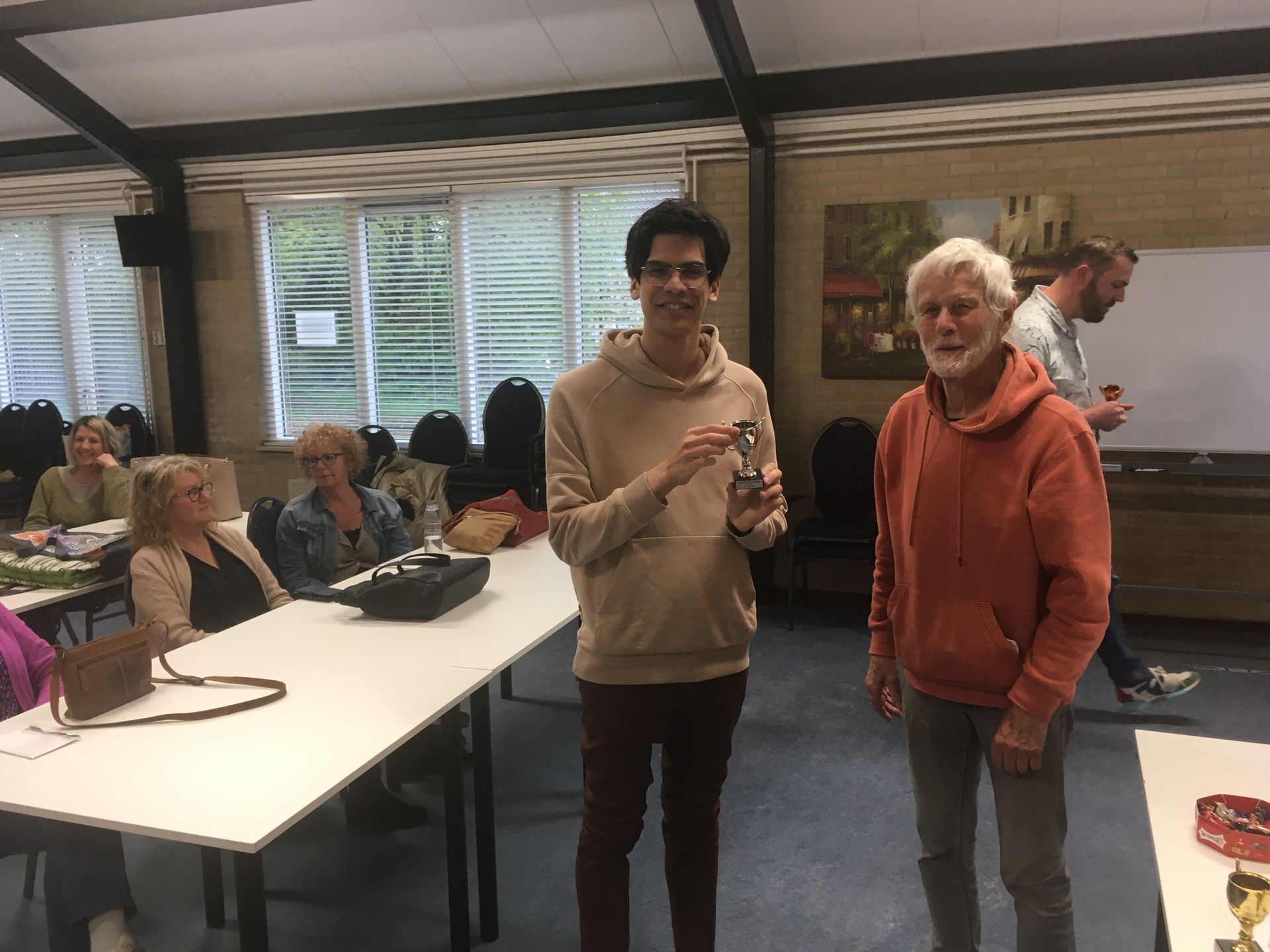 Een man in een beige hoodie houdt een trofee vast naast een oudere man in een oranje shirt; binnen zitten andere mensen aan tafels.