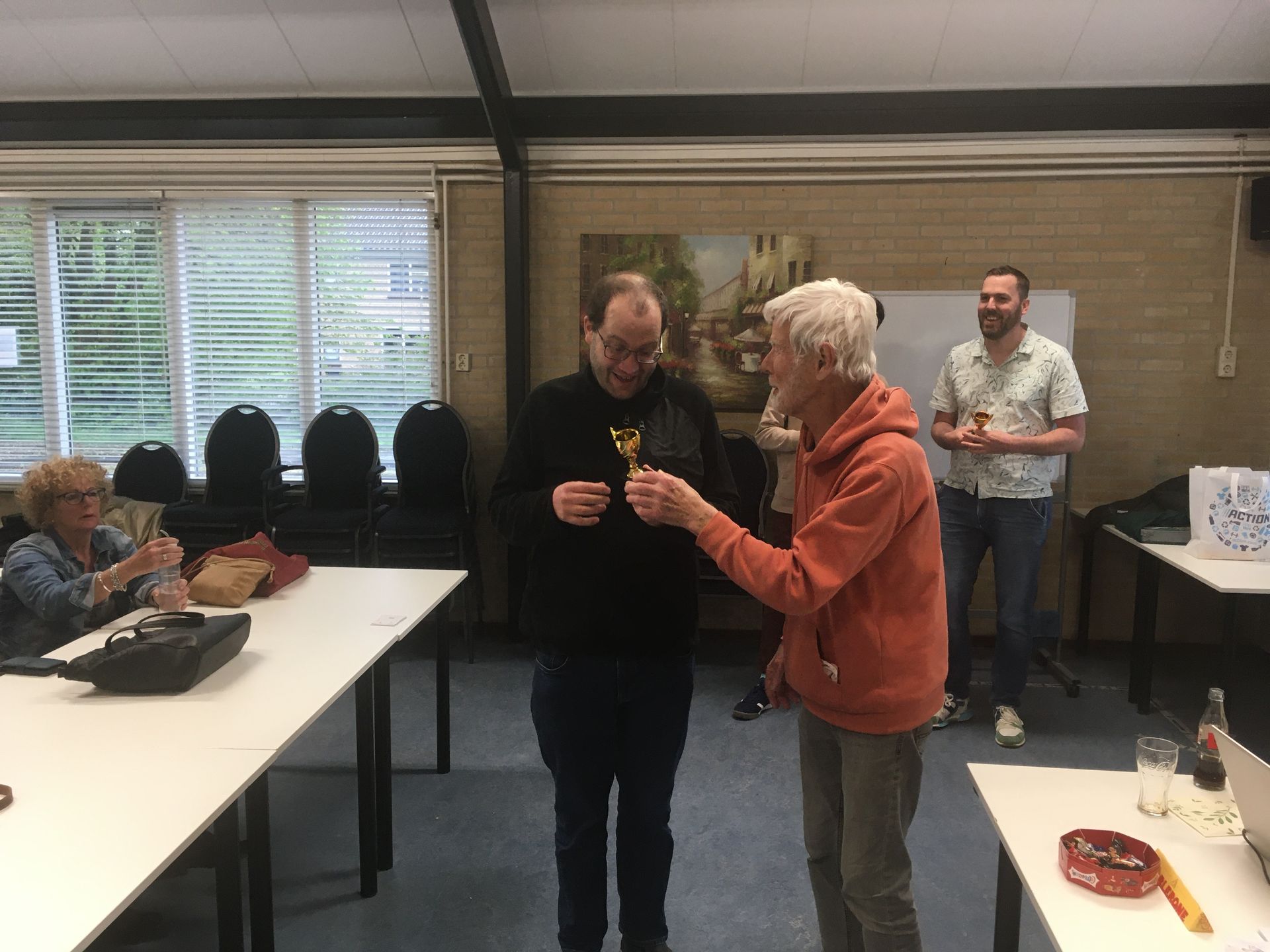 Een man in een zwart shirt ontvangt een trofee van een oudere man in een oranje hoodie, in een kamer. Andere mensen kijken toe.