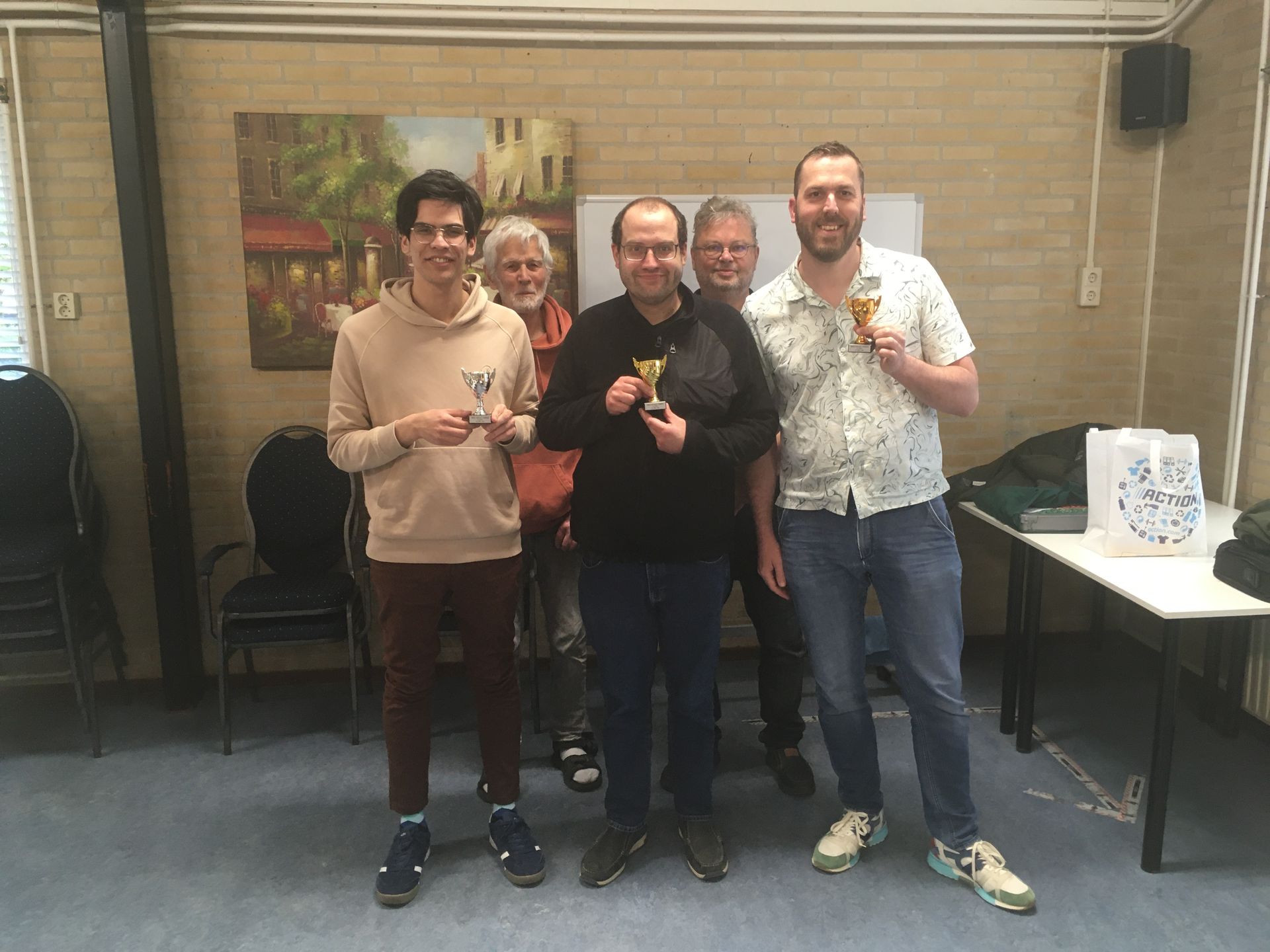 Een groep mensen met trofeeën in hun handen, lachend, binnen. Op de achtergrond zijn een schilderij en een tafel te zien.