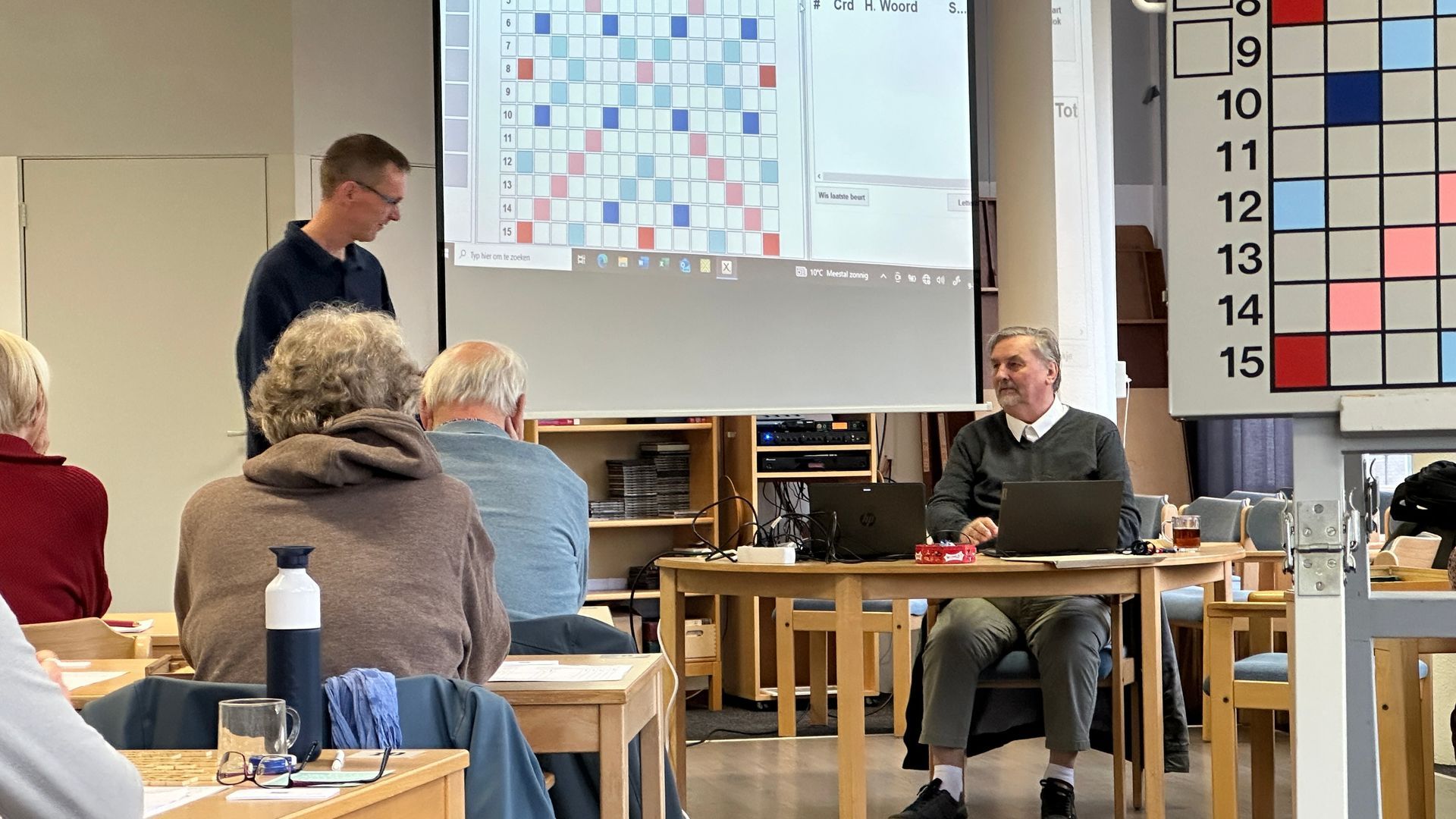 Een lezing in een zaal: een man spreekt terwijl een presentatie wordt getoond. Het publiek kijkt toe, met een spelbord ernaast.