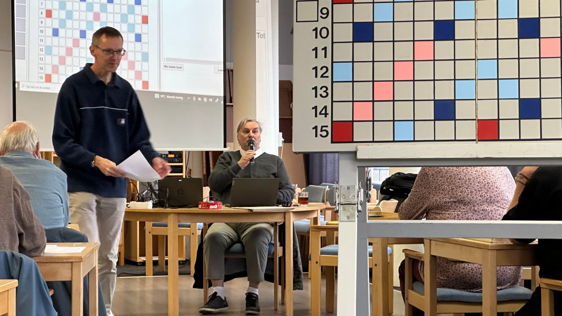 Een man geeft een presentatie tijdens een Scrabble-evenement met een bord, zittende deelnemers en een projector.