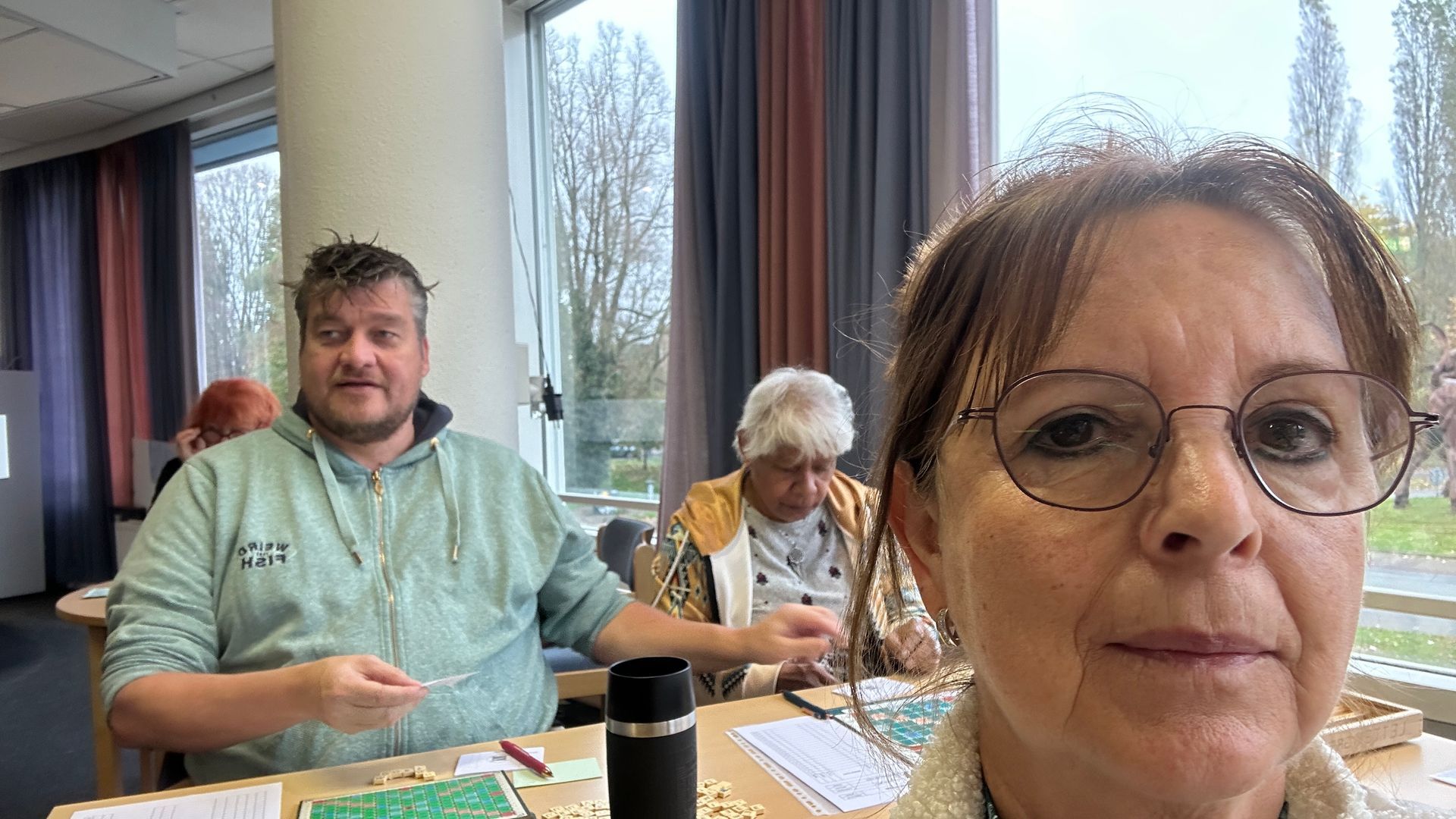 Een vrouw maakt een selfie in een kamer met twee andere mensen; ze spelen een spel. Op de achtergrond zijn ramen te zien.