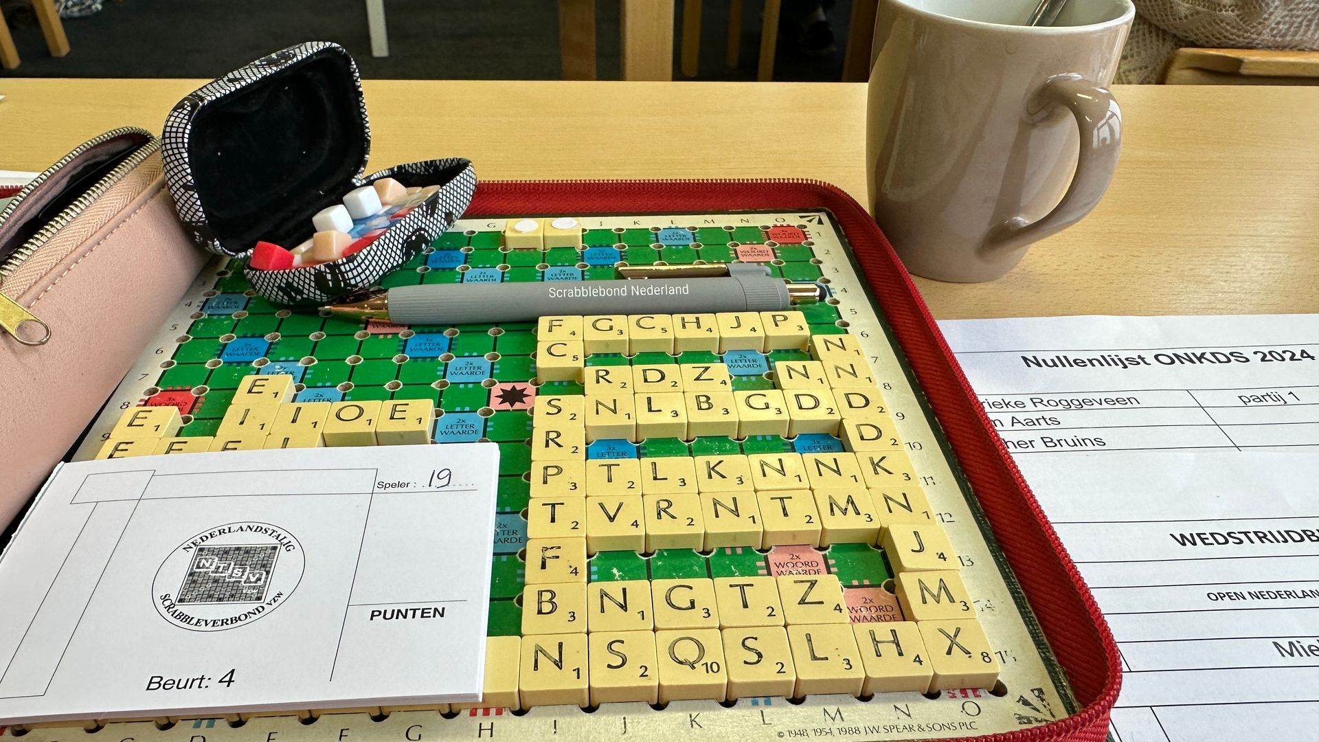 Een Scrabble-bord in gebruik op een tafel met een mok, etui, papier en pen.
