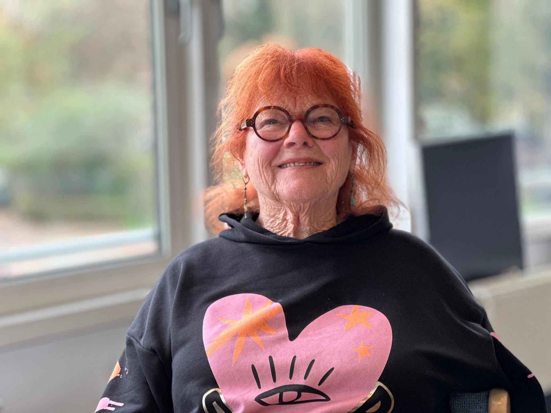 Een vrouw met rood haar en een bril lacht, terwijl ze een zwarte sweater met een roze hartje draagt.