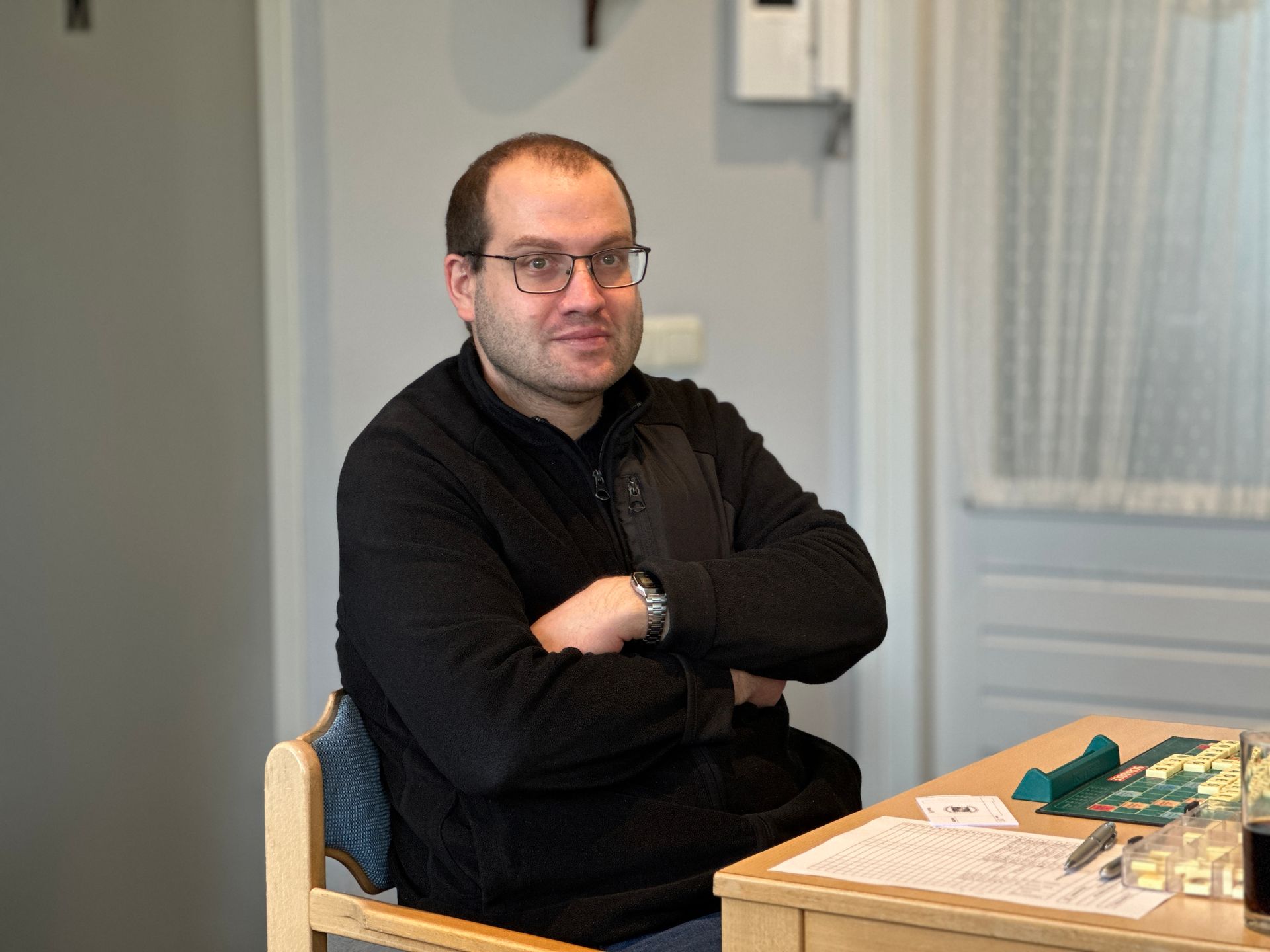 Een man met een bril zit aan een tafel, met zijn armen over elkaar, en glimlacht. Hij draagt ​​een zwarte trui. Binnen.