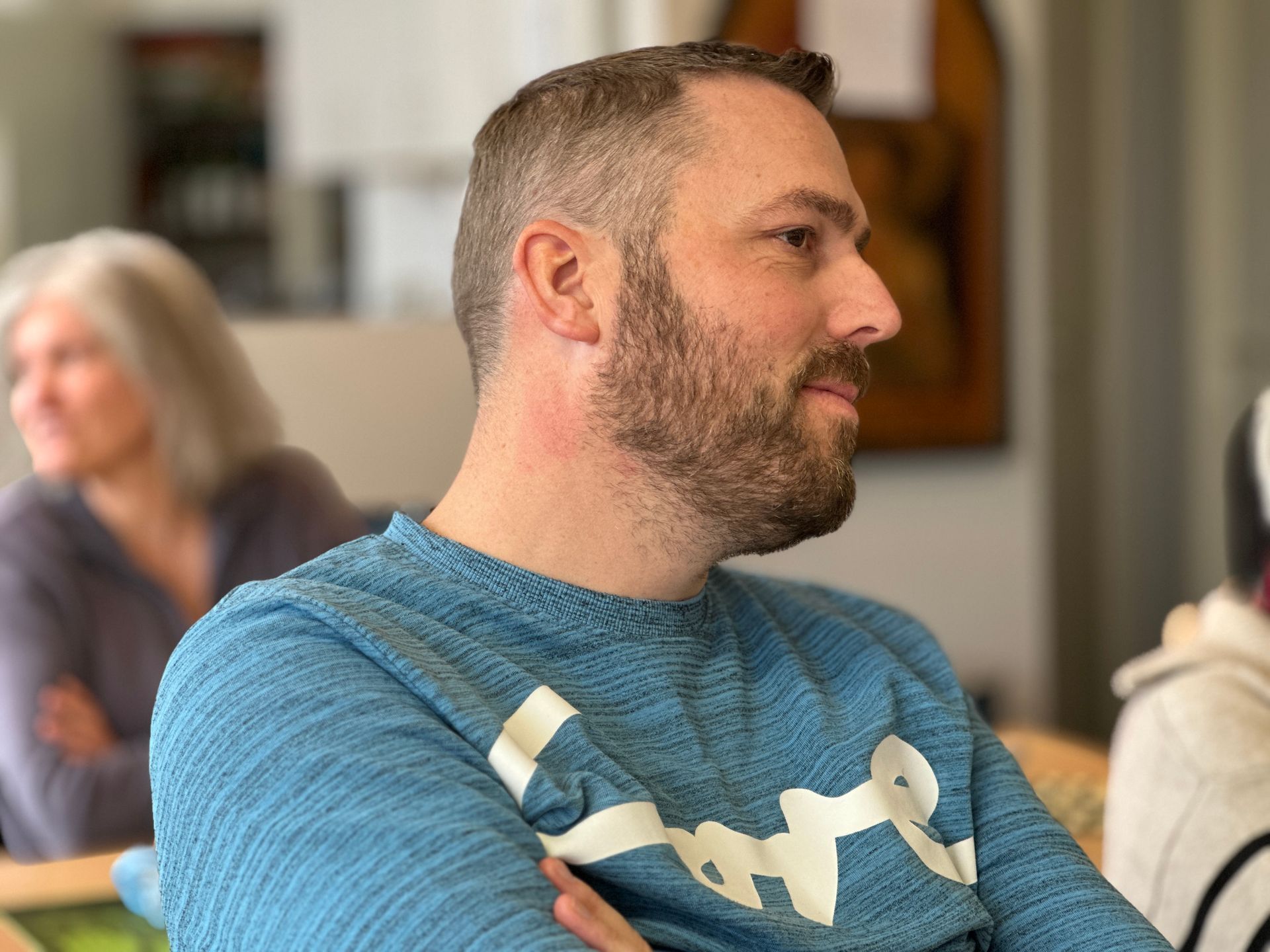 Een man met een baard, in een blauwe trui, zit binnen met zijn armen over elkaar en kijkt opzij.