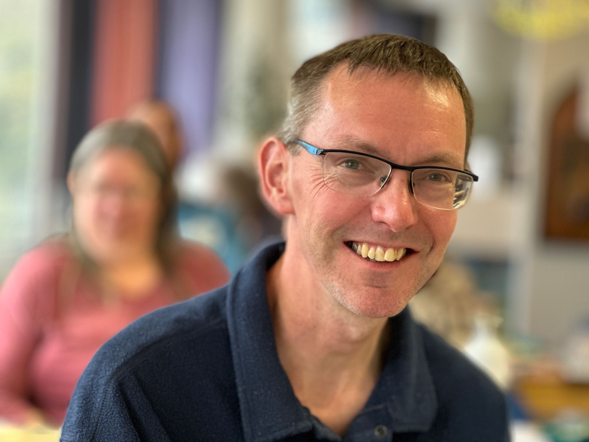 Een man met een bril lacht en draagt ​​een blauw shirt. Op de achtergrond is een vrouw in een roze shirt wazig te zien.