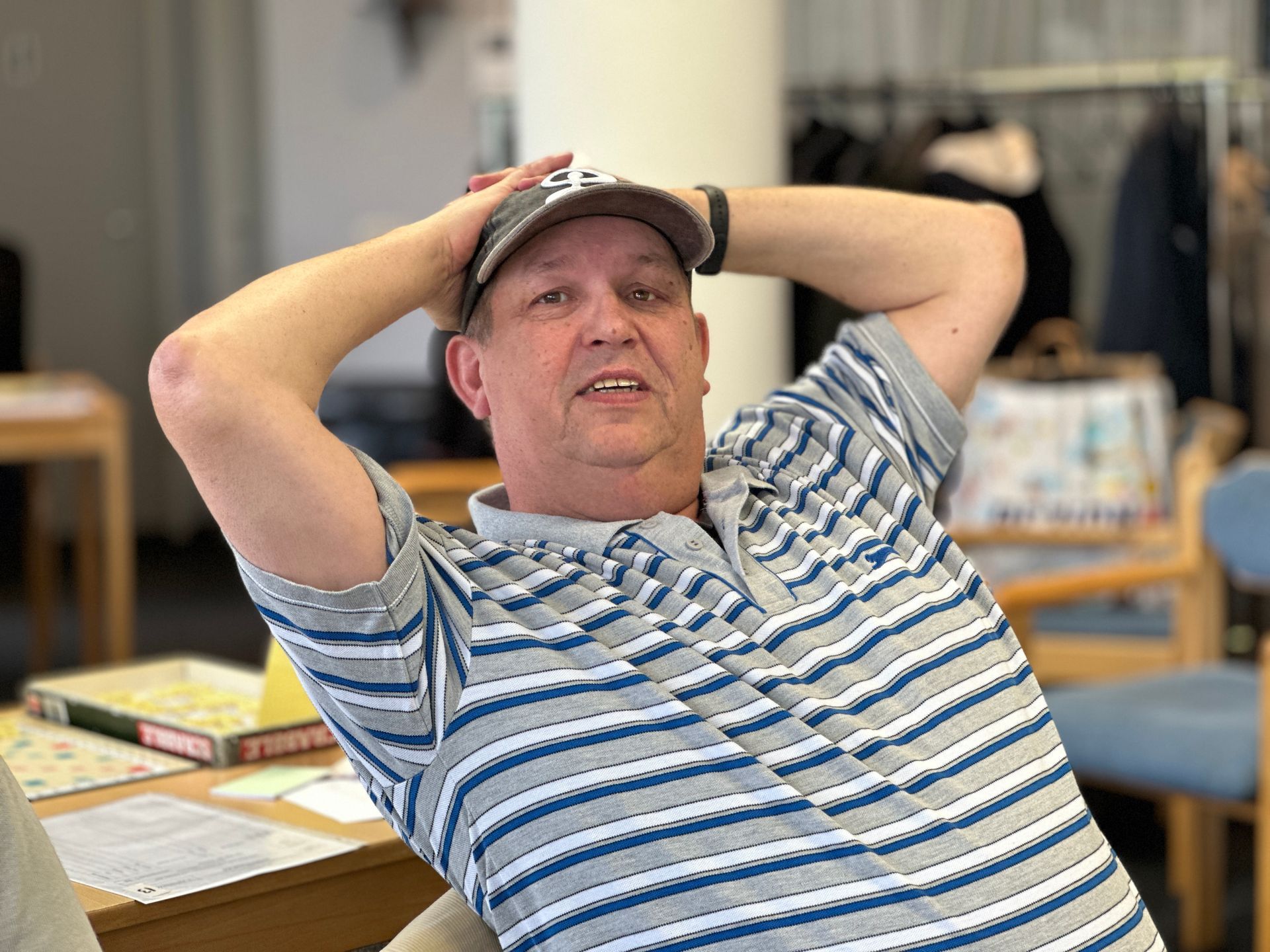 Een man in een gestreept shirt en een baseballpet laat zijn armen achter zijn hoofd rusten, binnenshuis.