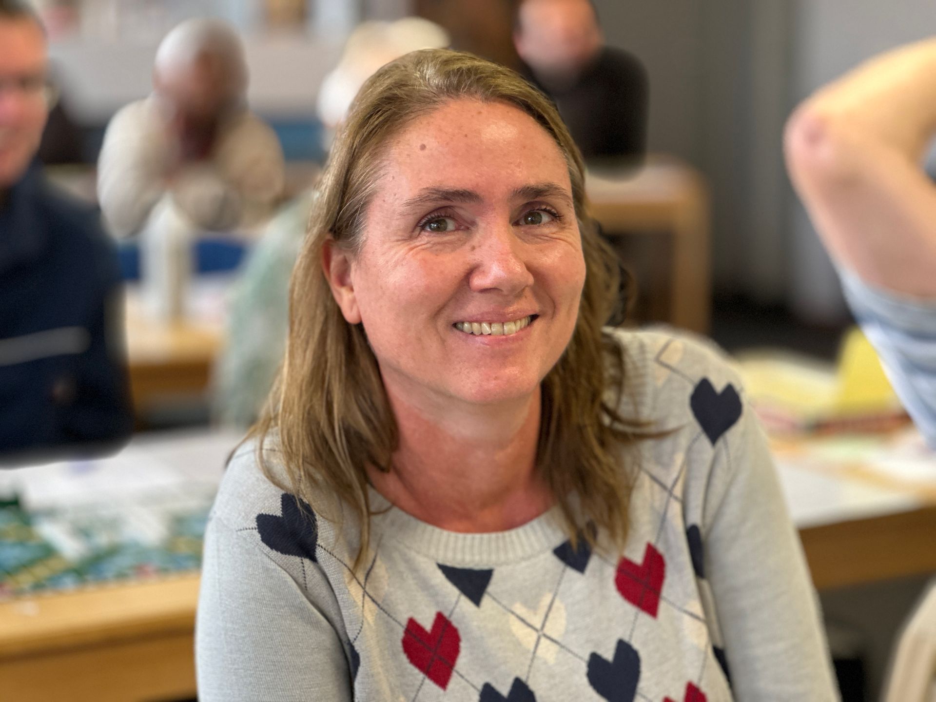 Een vrouw in een trui lacht naar de camera; op de achtergrond zijn andere mensen aan een tafel te zien.