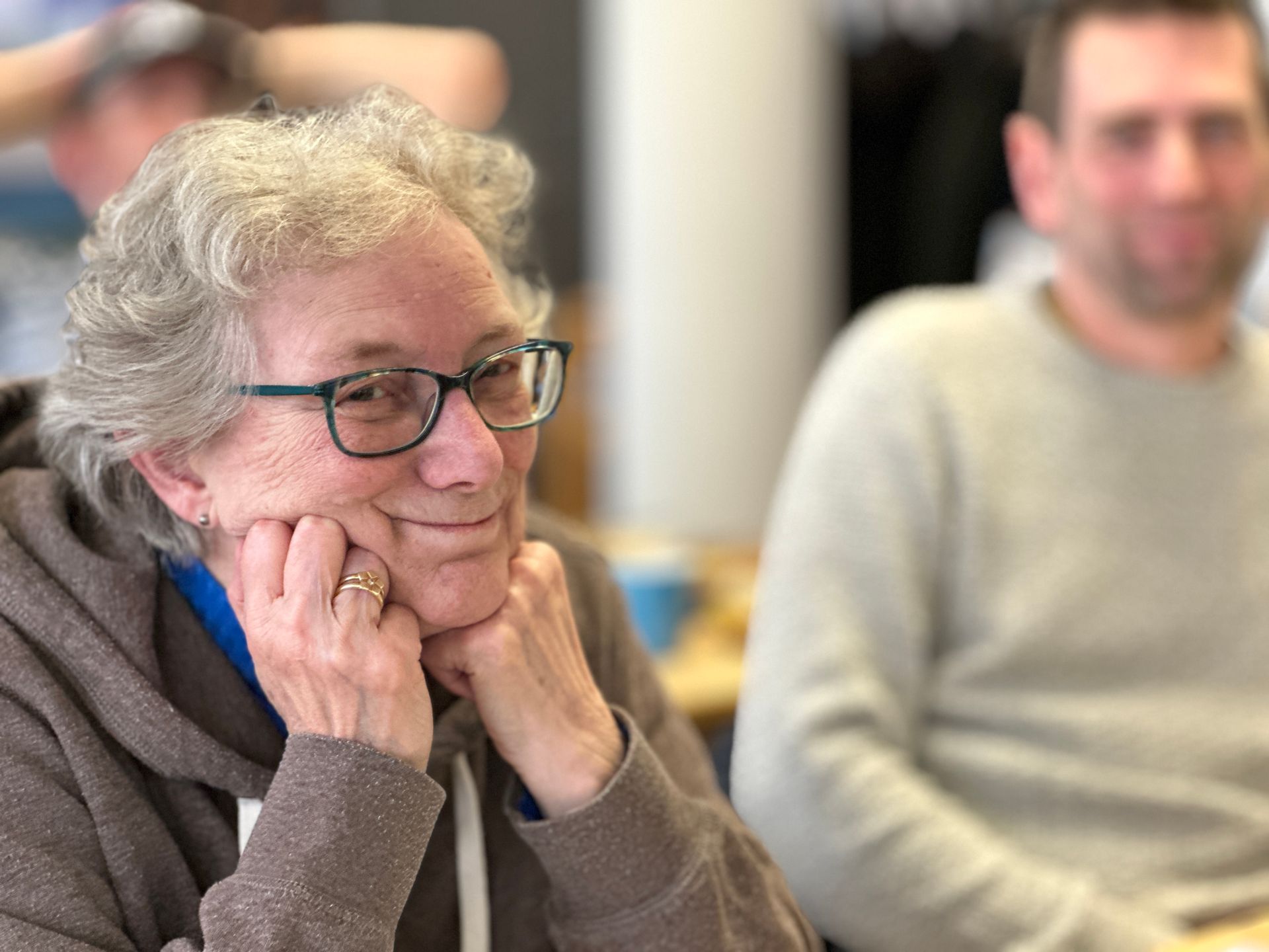 Vrouw met bril, kin rustend op handen, glimlachend; man op de achtergrond, wazig.