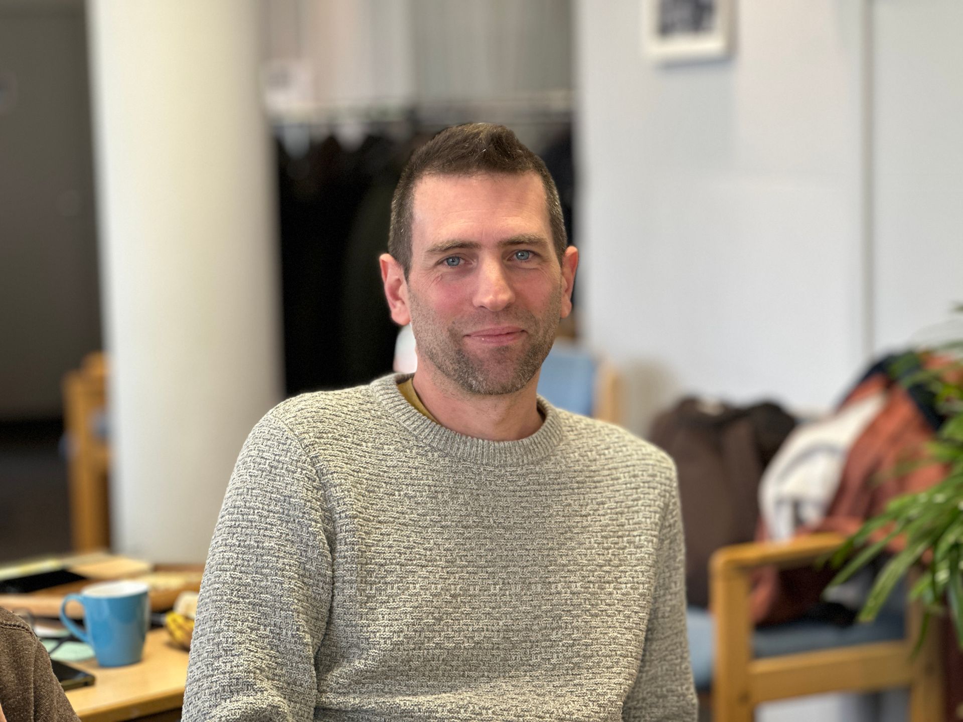 Een man in een grijze trui glimlacht, zittend in een kamer. Op de achtergrond zijn objecten en een pilaar wazig.