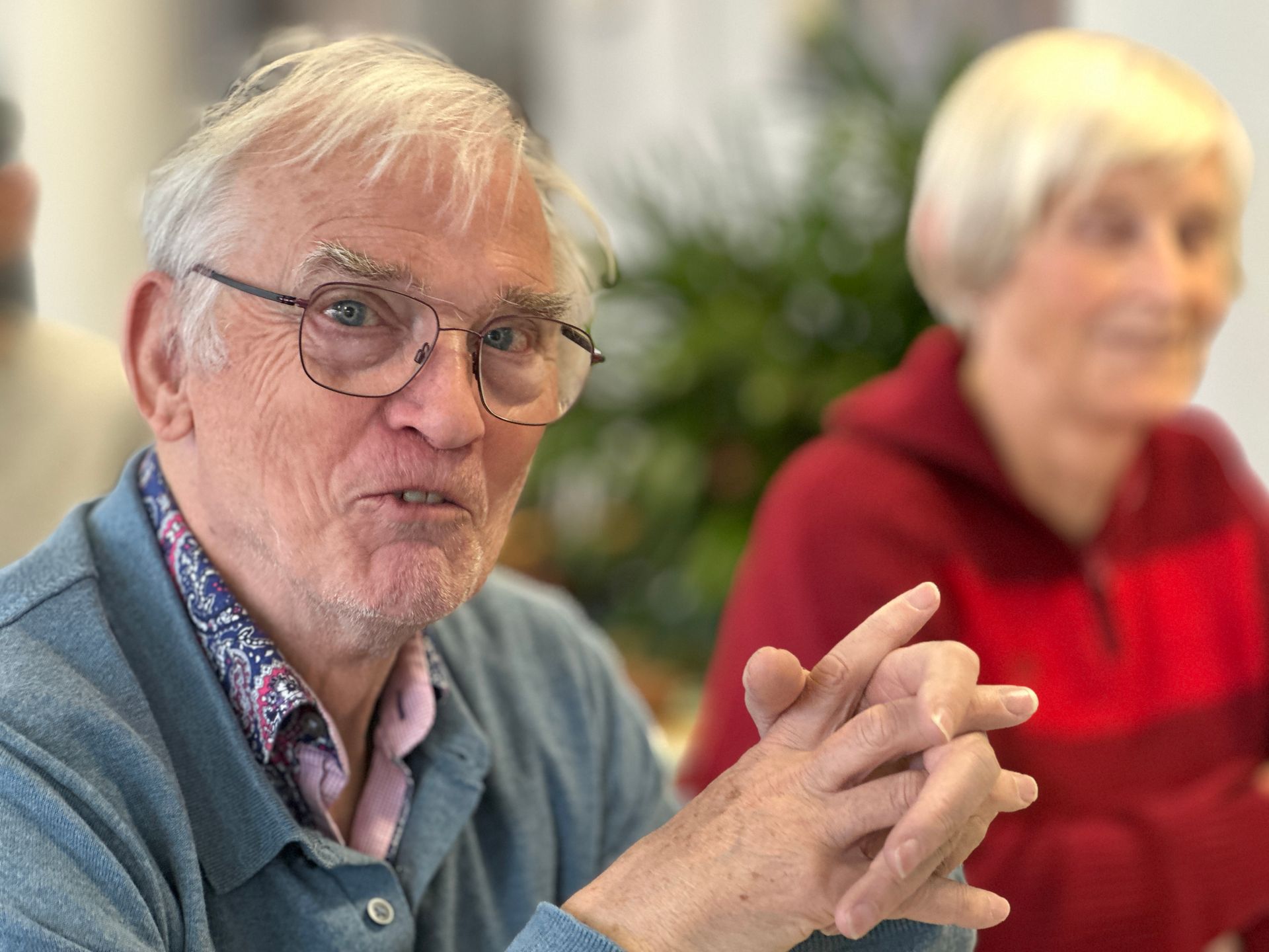 Oudere man met bril, blauwe trui, sprekend met gevouwen handen; een andere persoon is wazig op de achtergrond te zien.