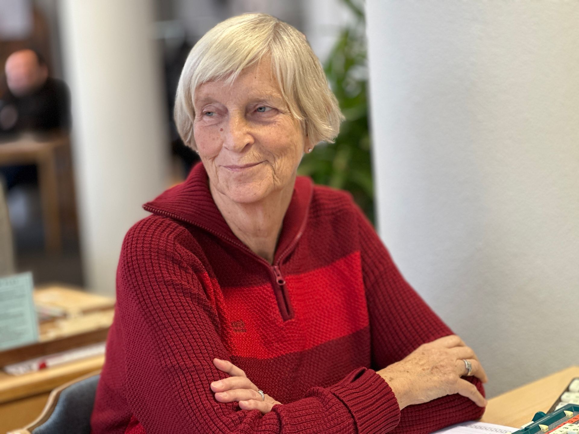 Een vrouw met kort blond haar en een rode trui zit glimlachend aan een tafel.