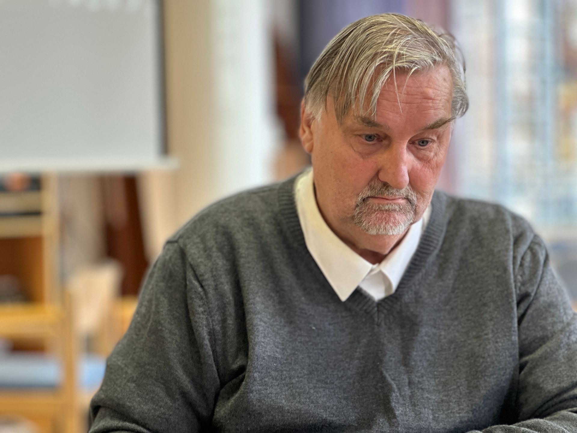 Een man in een grijze trui en een wit overhemd kijkt naar beneden, binnen.