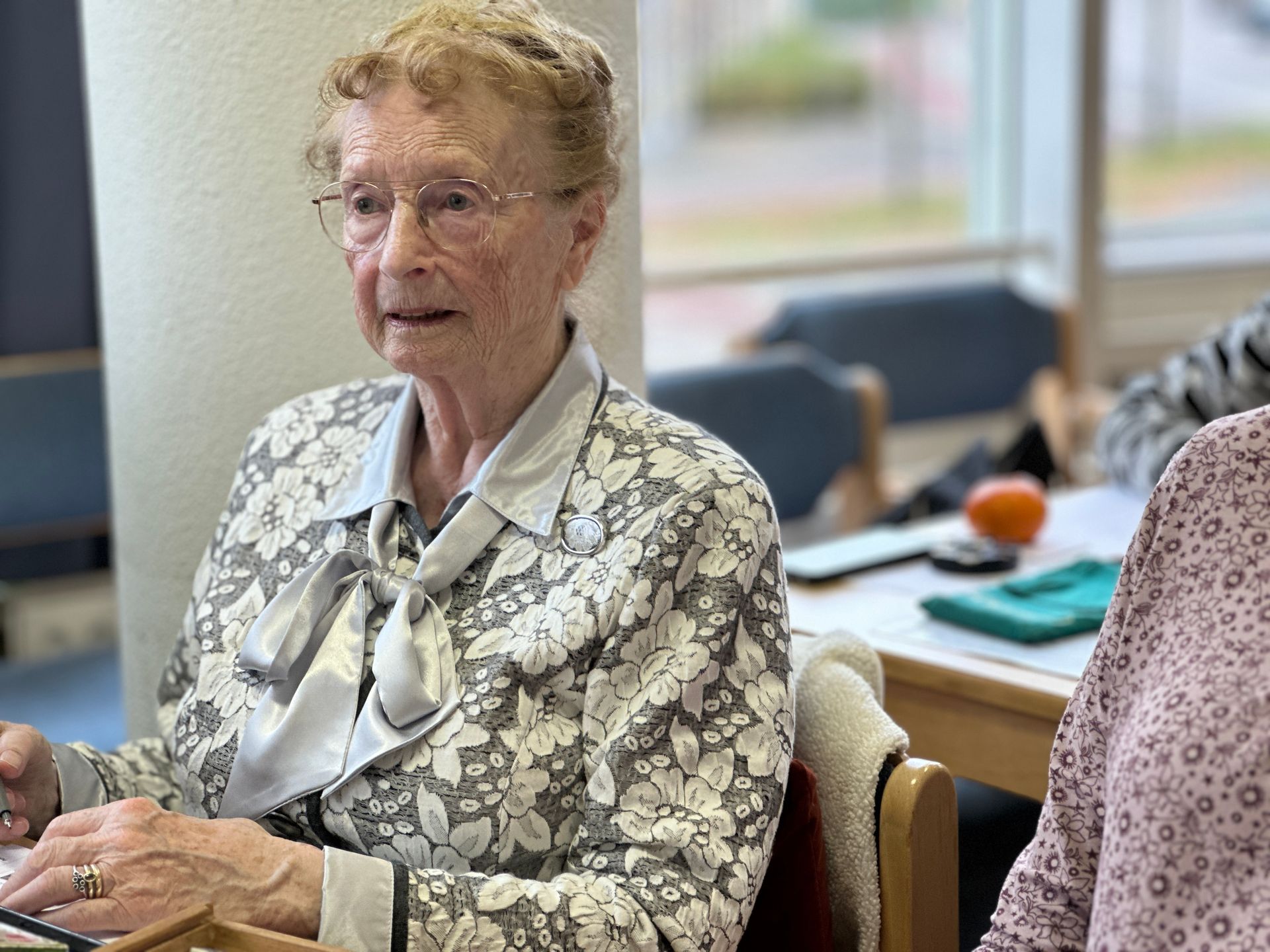 Een oudere vrouw met een bril en een blouse met bloemenprint zit aan een tafel en kijkt voor zich uit.