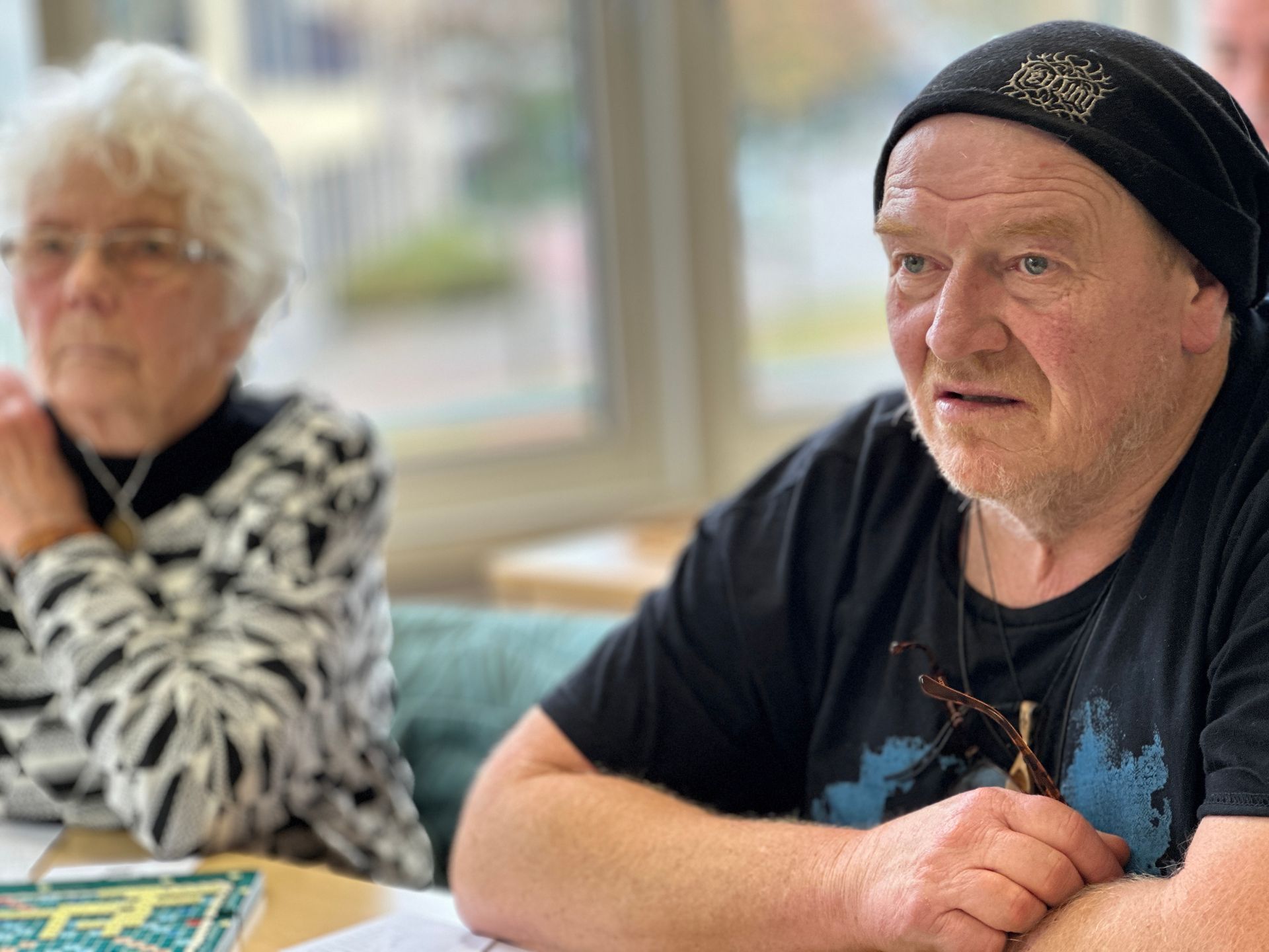 Een man met een zwarte pet en een T-shirt spreekt. Op de achtergrond is een vrouw te zien met een bril en een top met een patroon.
