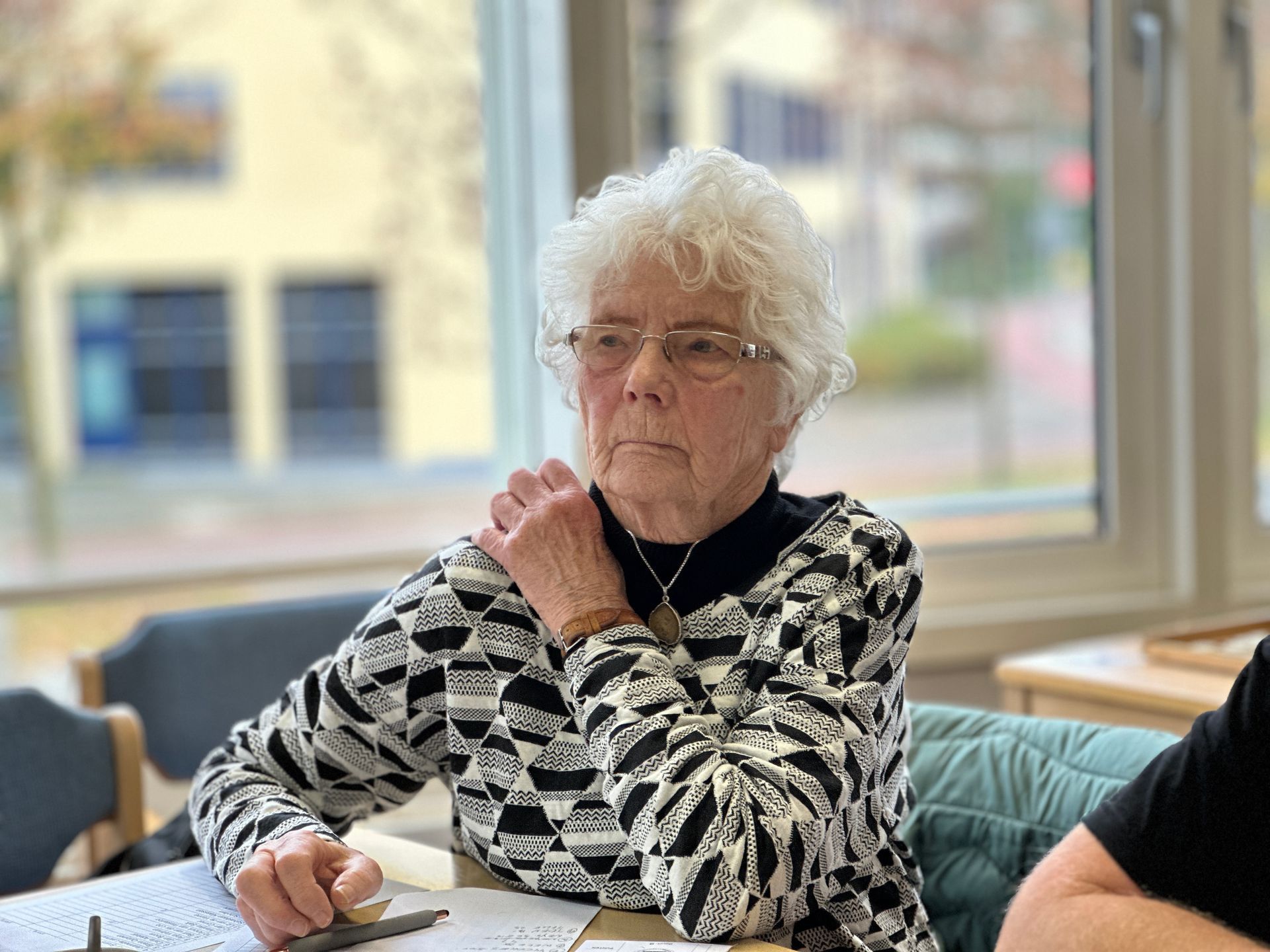 Vrouw met wit haar, een bril en een zwart-wit gestreept shirt, zit bij het raam.