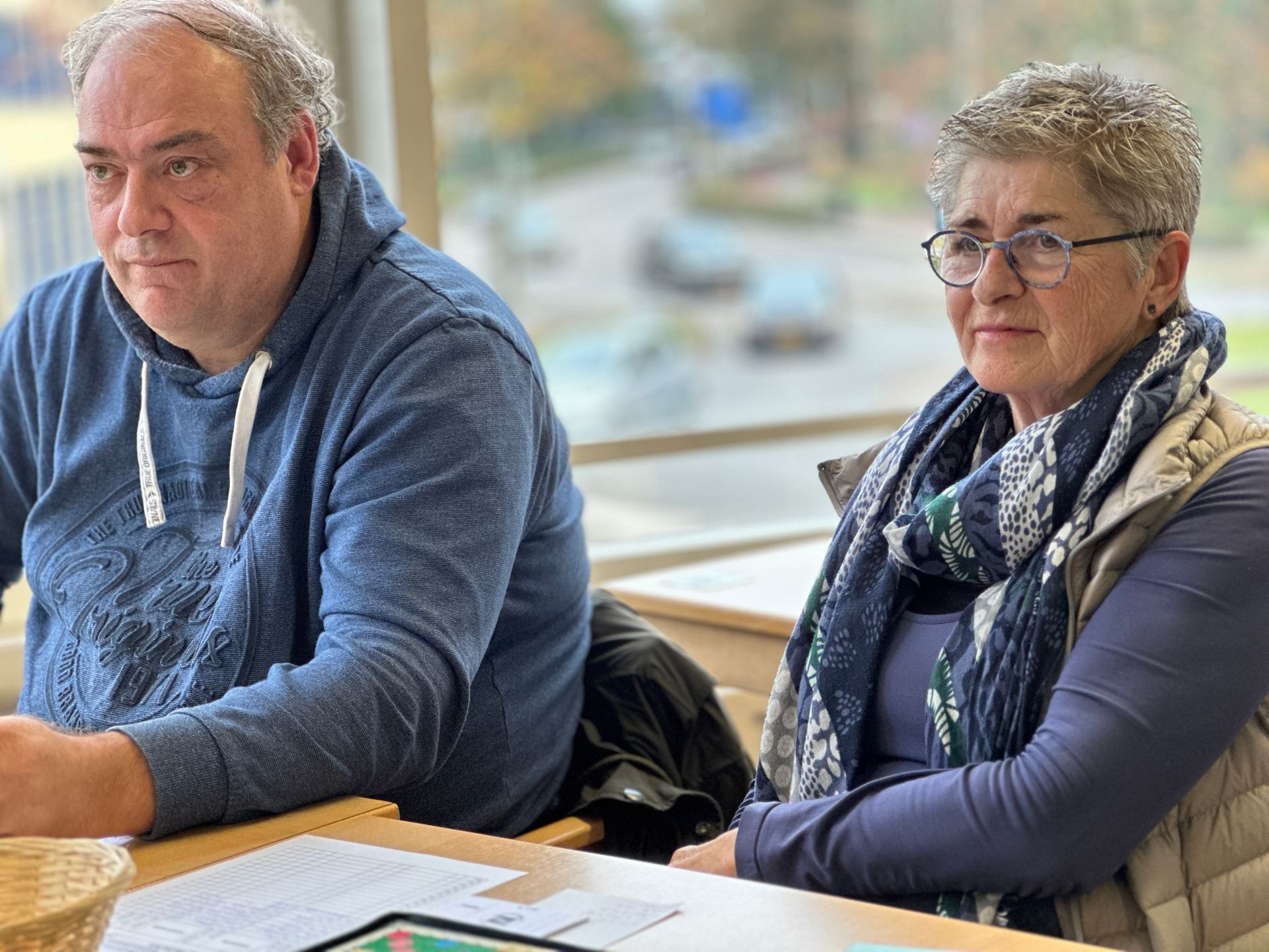 Een man in een blauwe hoodie en een vrouw met een bril zitten aan een tafel en kijken opzij, binnen.