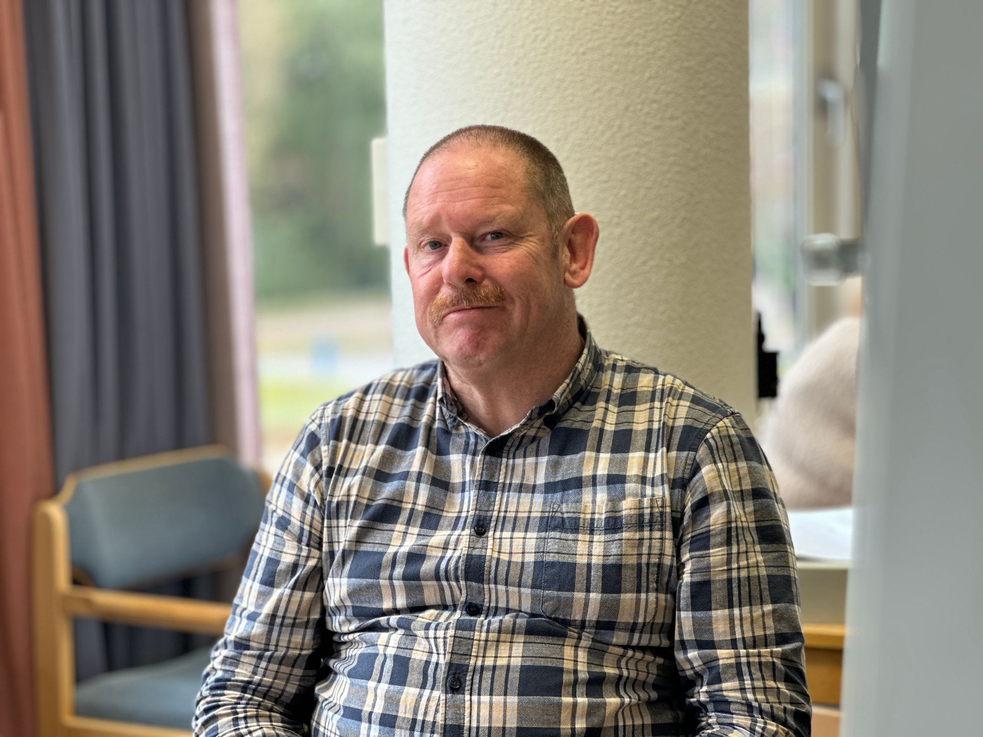 Een man met een geruit overhemd zit binnen en kijkt naar de camera.