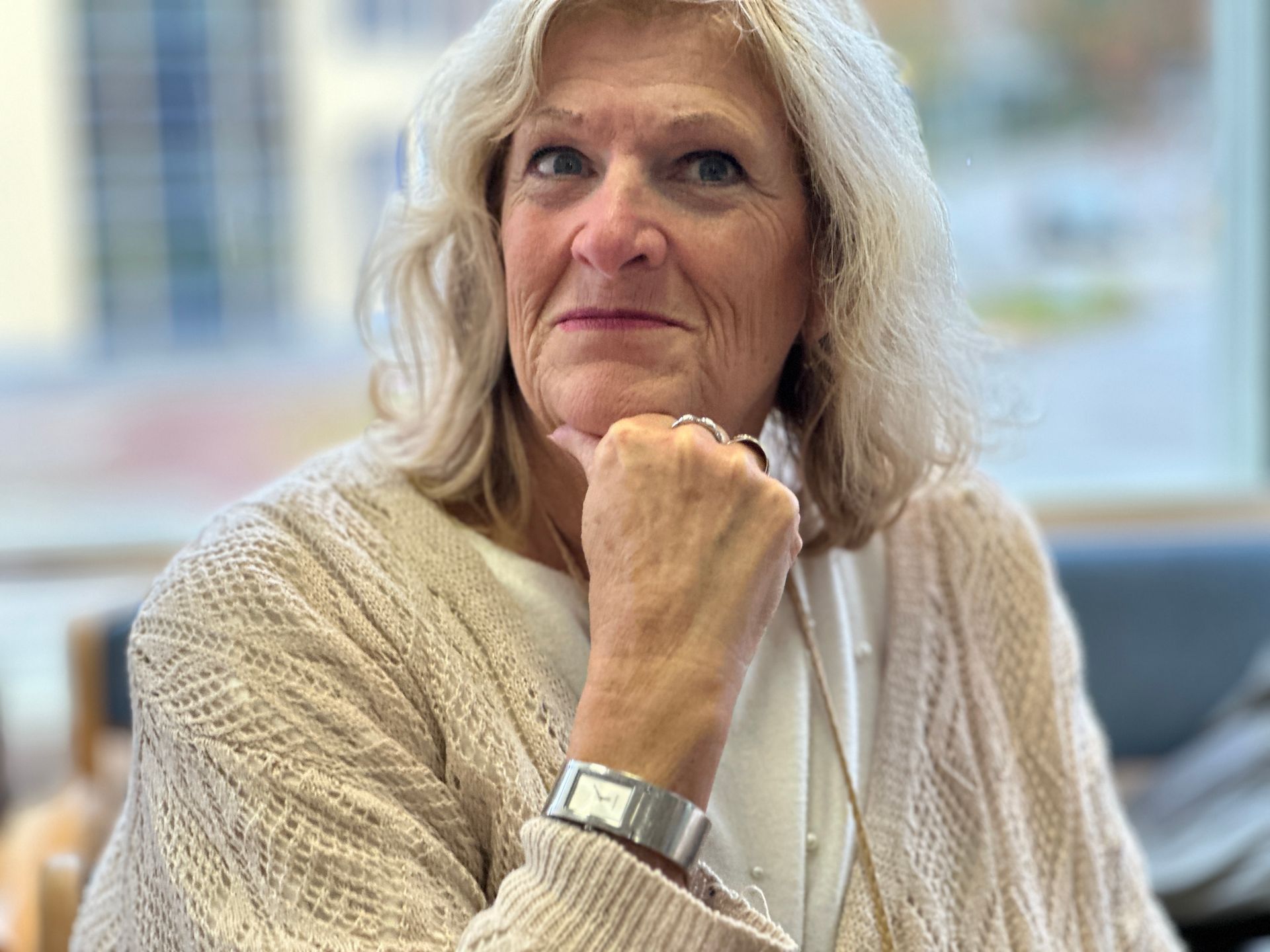Vrouw met blond haar, kin rustend op hand, gekleed in een crèmekleurige trui en zilveren armband, kijkt in de camera.