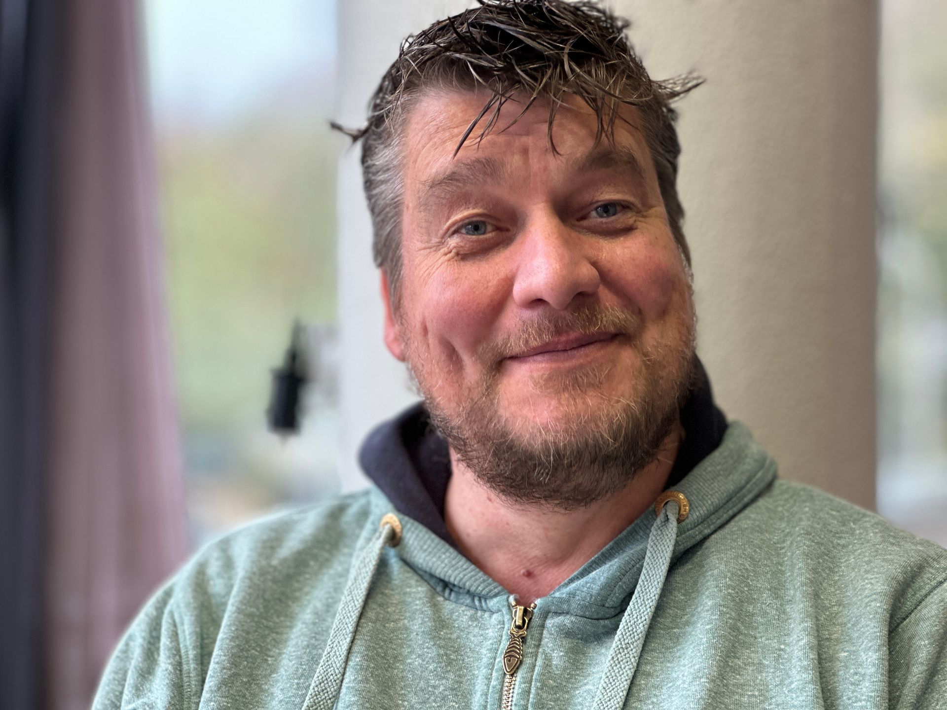 Man met lichtbruin haar en een baard, gekleed in een lichtblauwe hoodie, glimlacht lichtjes binnenshuis.