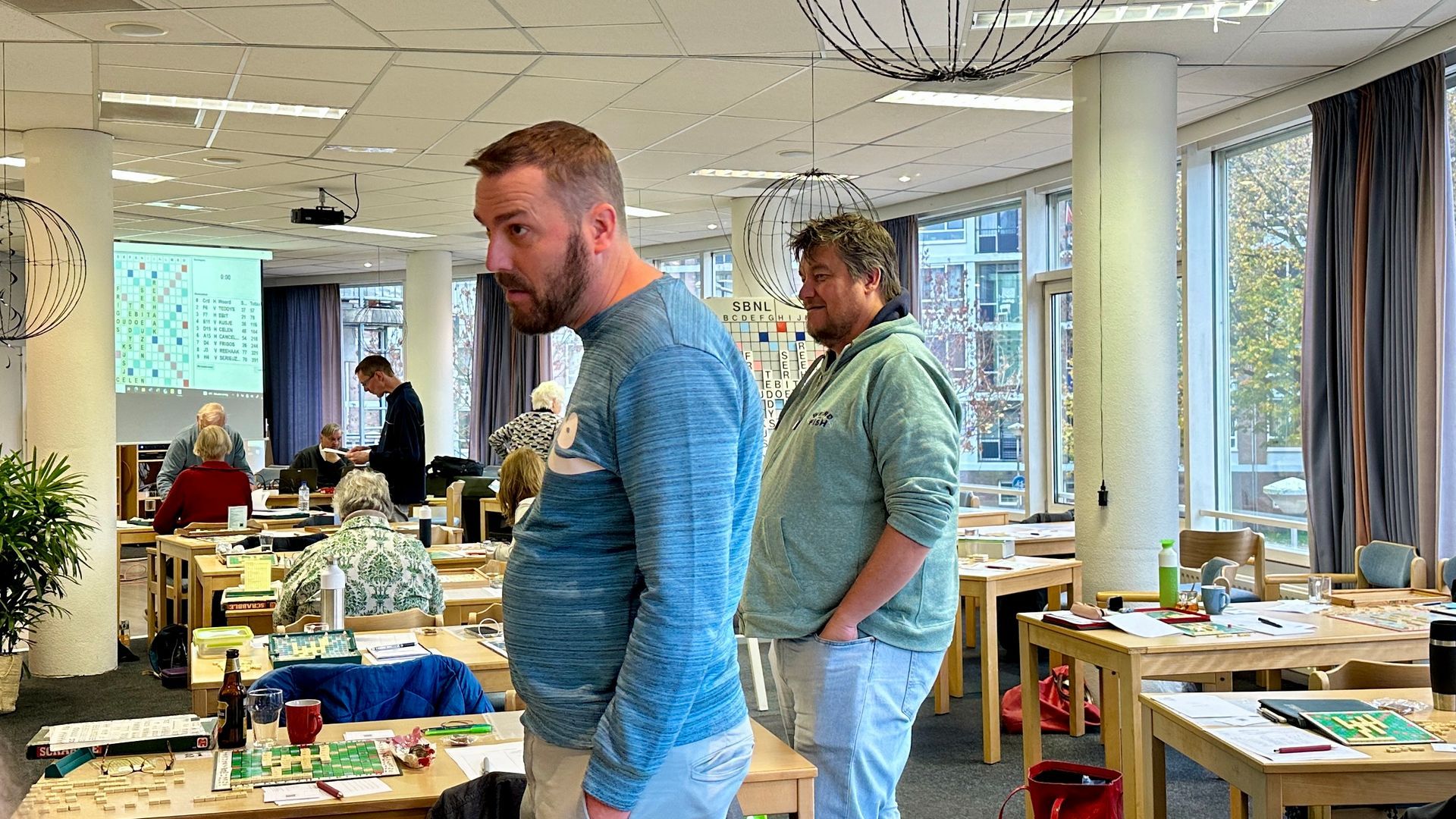 Twee mannen in een kantooromgeving, de ene kijkt opzij, de andere iets anders. Tafels en een presentatie zijn zichtbaar.