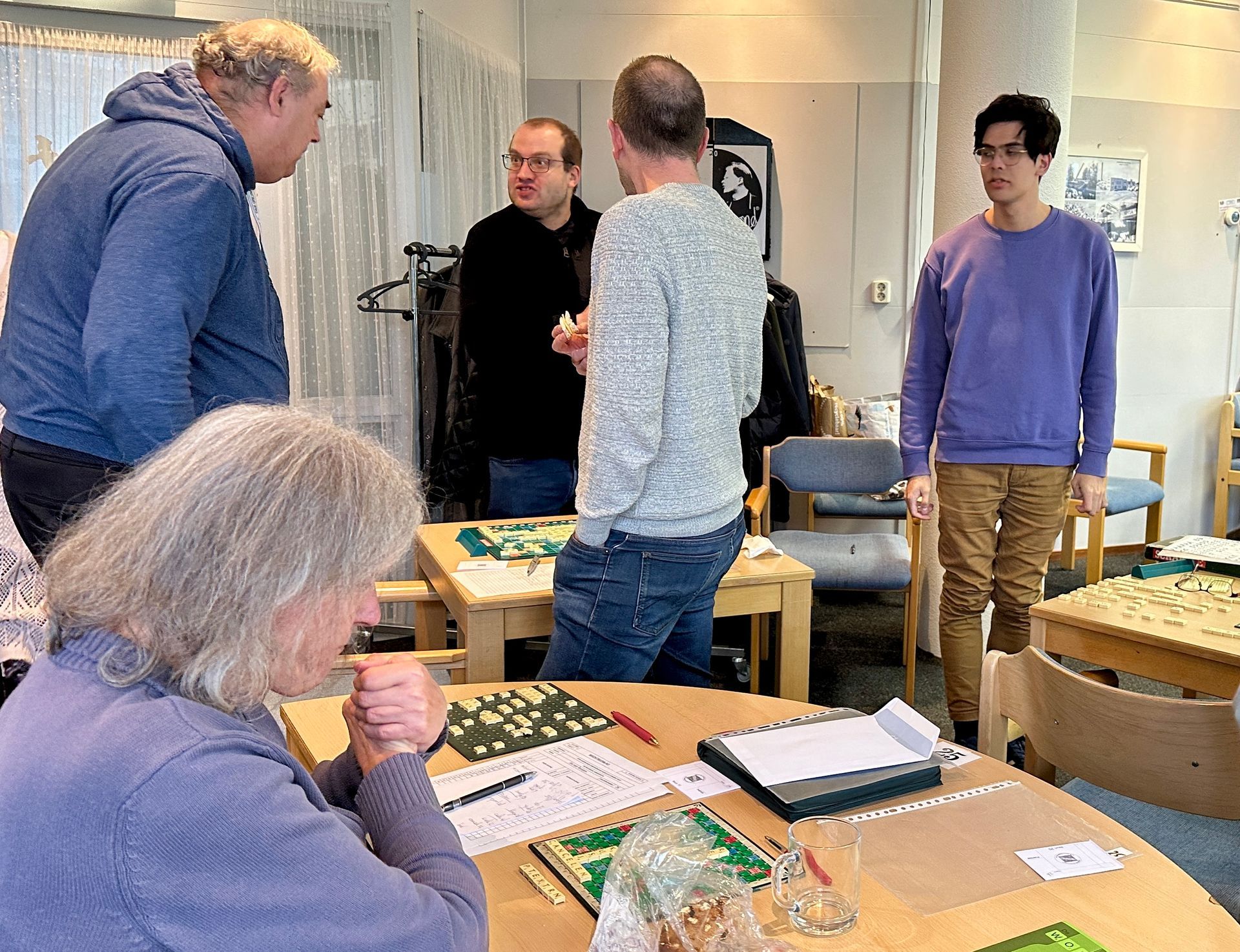 Mensen spelen een bordspel in een kamer, sommigen kijken naar anderen.