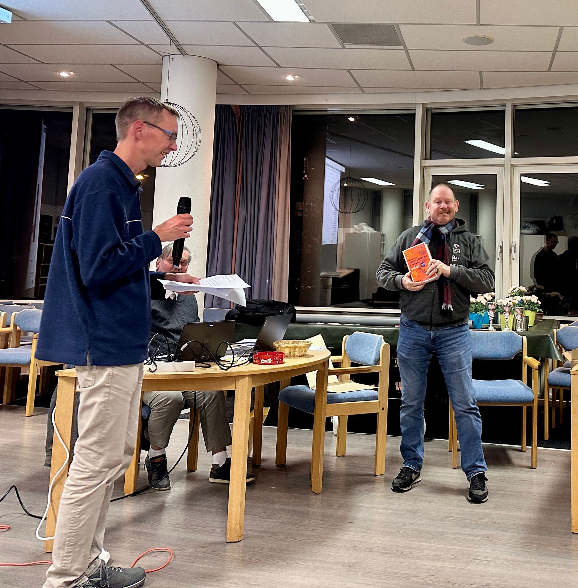 Een man spreekt in een microfoon, een andere man houdt een boek vast, beiden in een kamer met stoelen en een tafel.