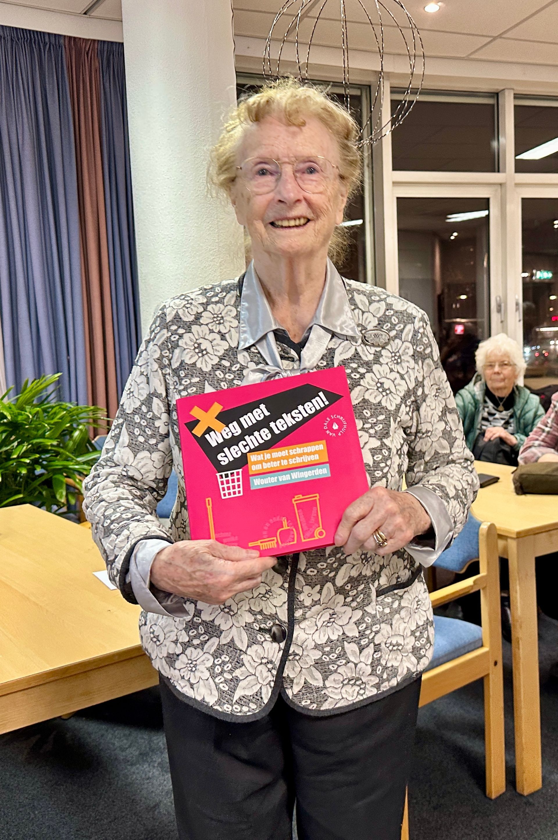 Vrouw met een roze boek in haar handen, glimlachend. Ze draagt ​​een jasje met bloemenprint en een bril. Binnen, bij een tafel.