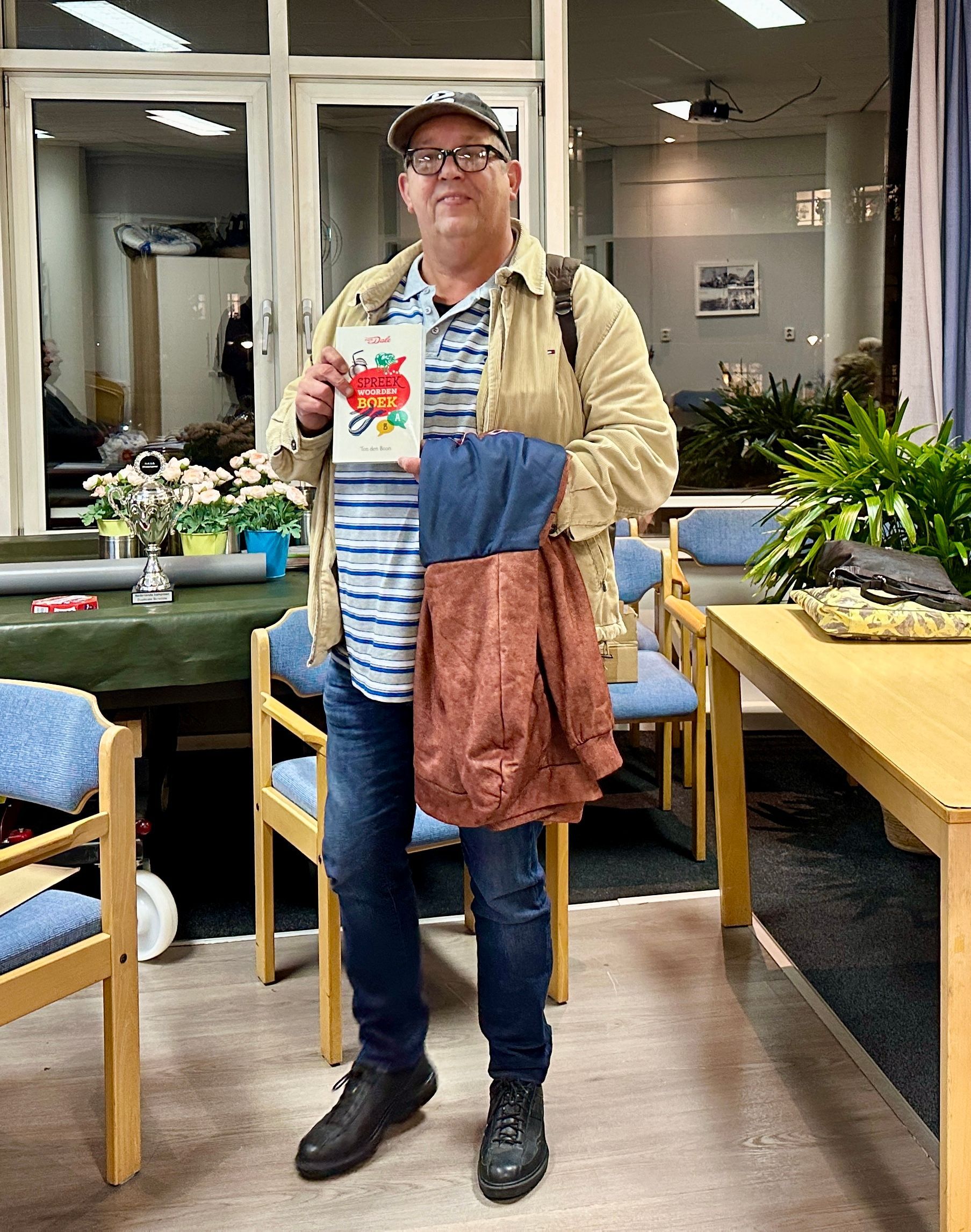 Een man met een boek en tassen staat in een kamer met tafels en stoelen.