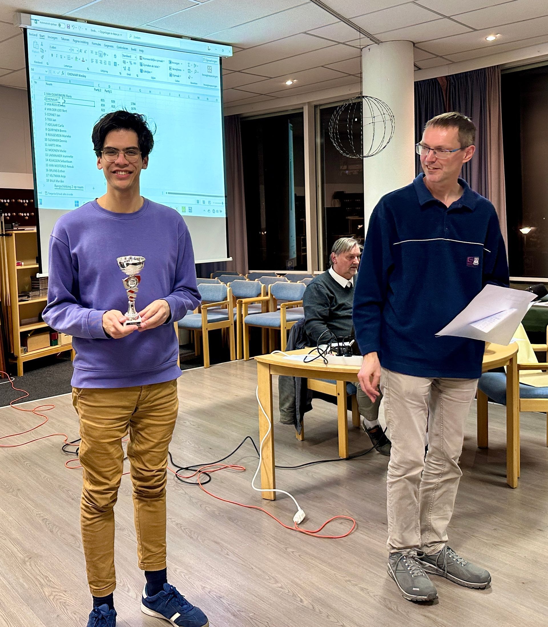 Een man in een paars pak houdt een trofee vast, een andere man heeft papieren in zijn handen. Ze staan ​​in een kamer met een projectiescherm en stoelen.