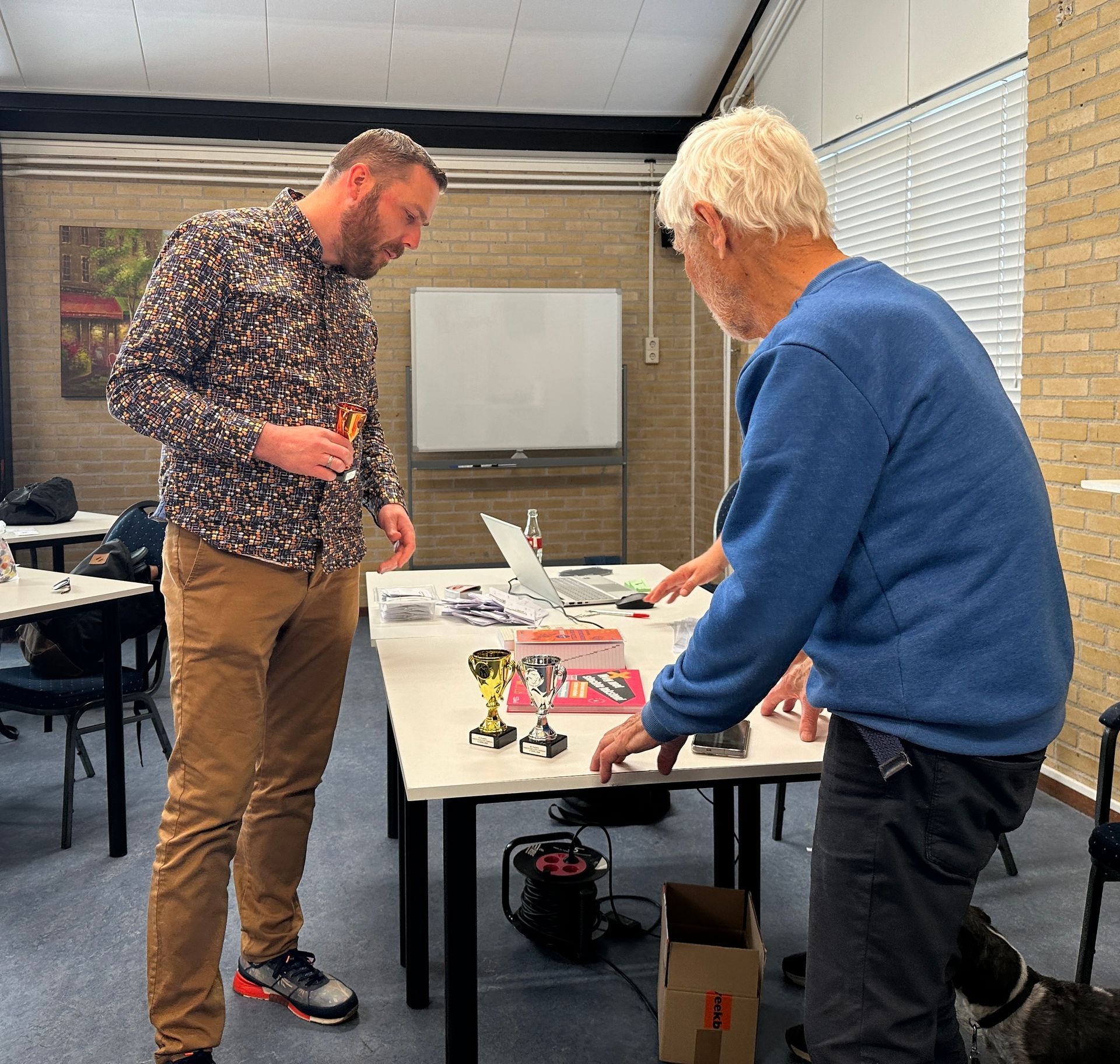 Twee mannen zitten aan een tafel met gereedschap en een laptop en lijken samen te werken.